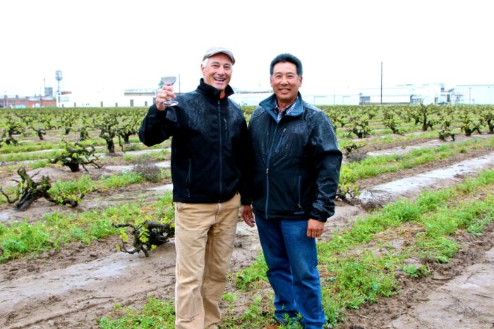 Two men in rain jackets standing in a vineyard, smiling, with grapevines and overcast sky in the background.