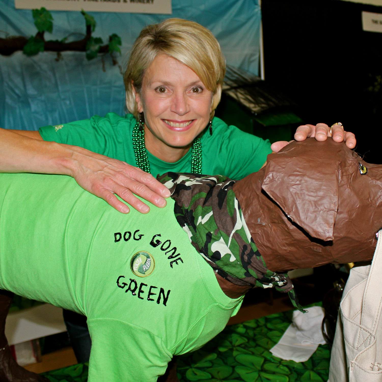 Woman smiling and hugging a dog dressed in a green shirt with 'Dog Gone Green' logo at Lodi Winegrape event.