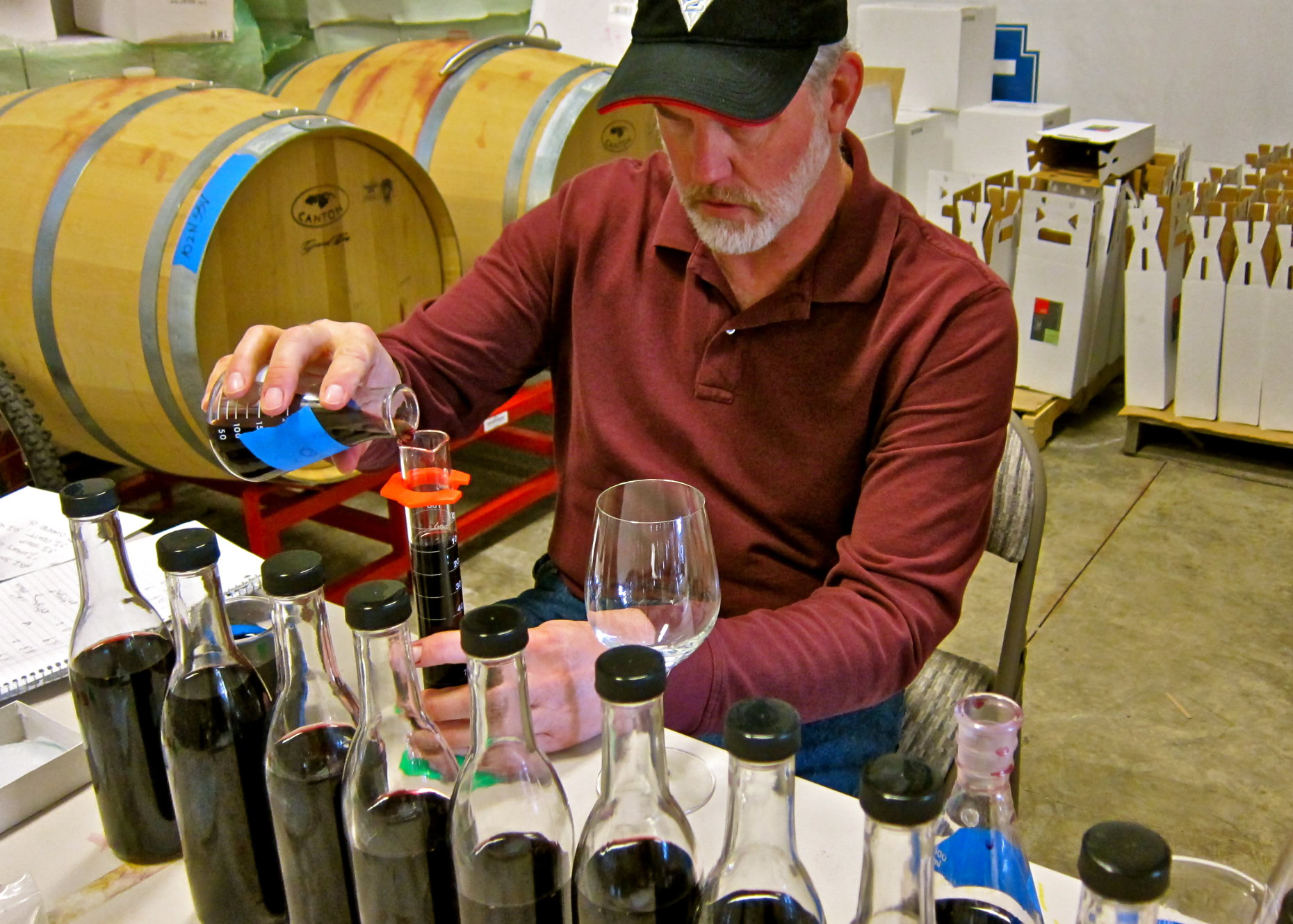 Man tasting wine samples at a wine tasting event with wine bottles and barrels in the background.