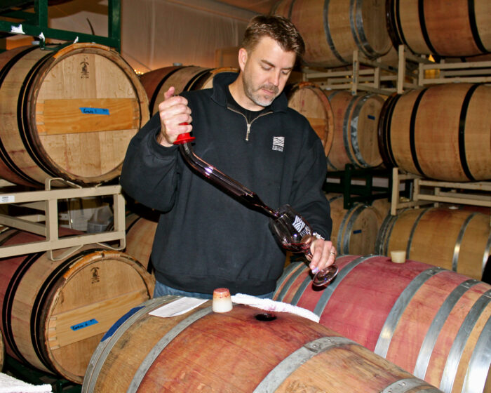 Winemaker pouring wine into a glass from a barrel in a wine cellar with stacked barrels in the background.