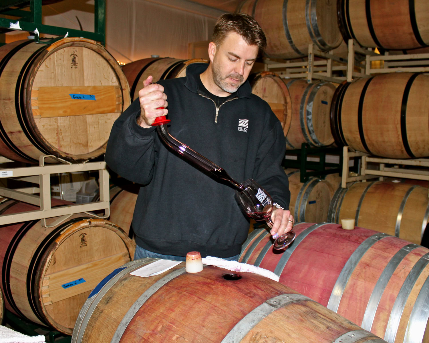 Winemaker pouring wine into a glass from a barrel in a wine cellar with stacked barrels in the background.