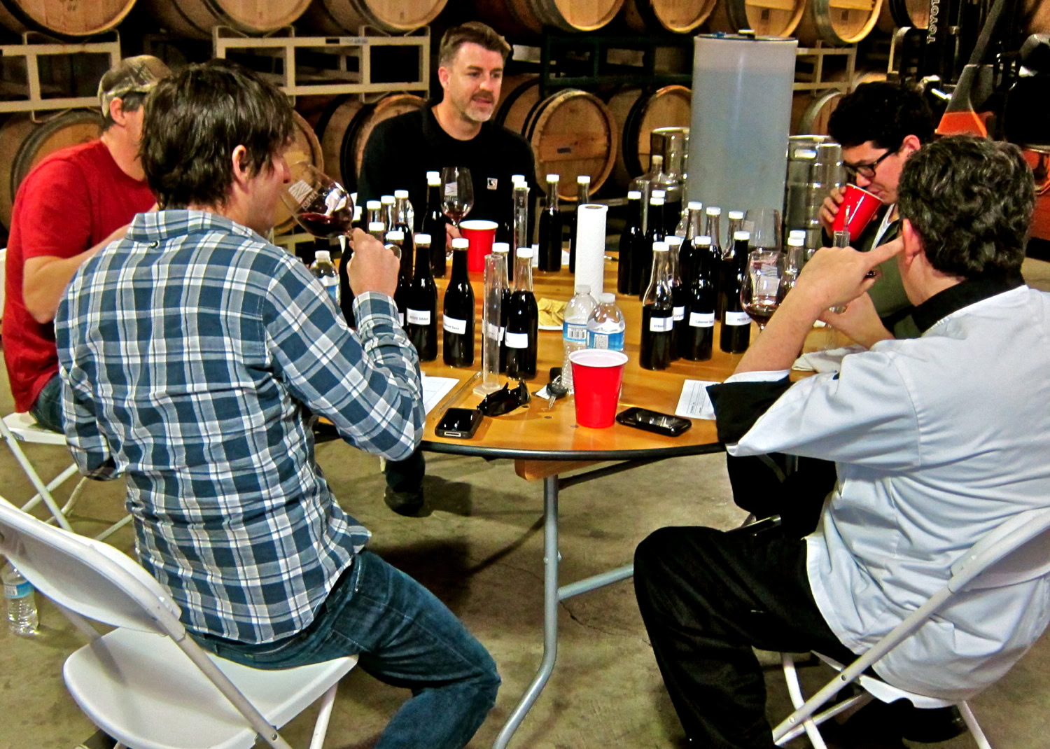 Group of people tasting wine at a winery, surrounded by wine bottles and barrels, in a wine production setting.