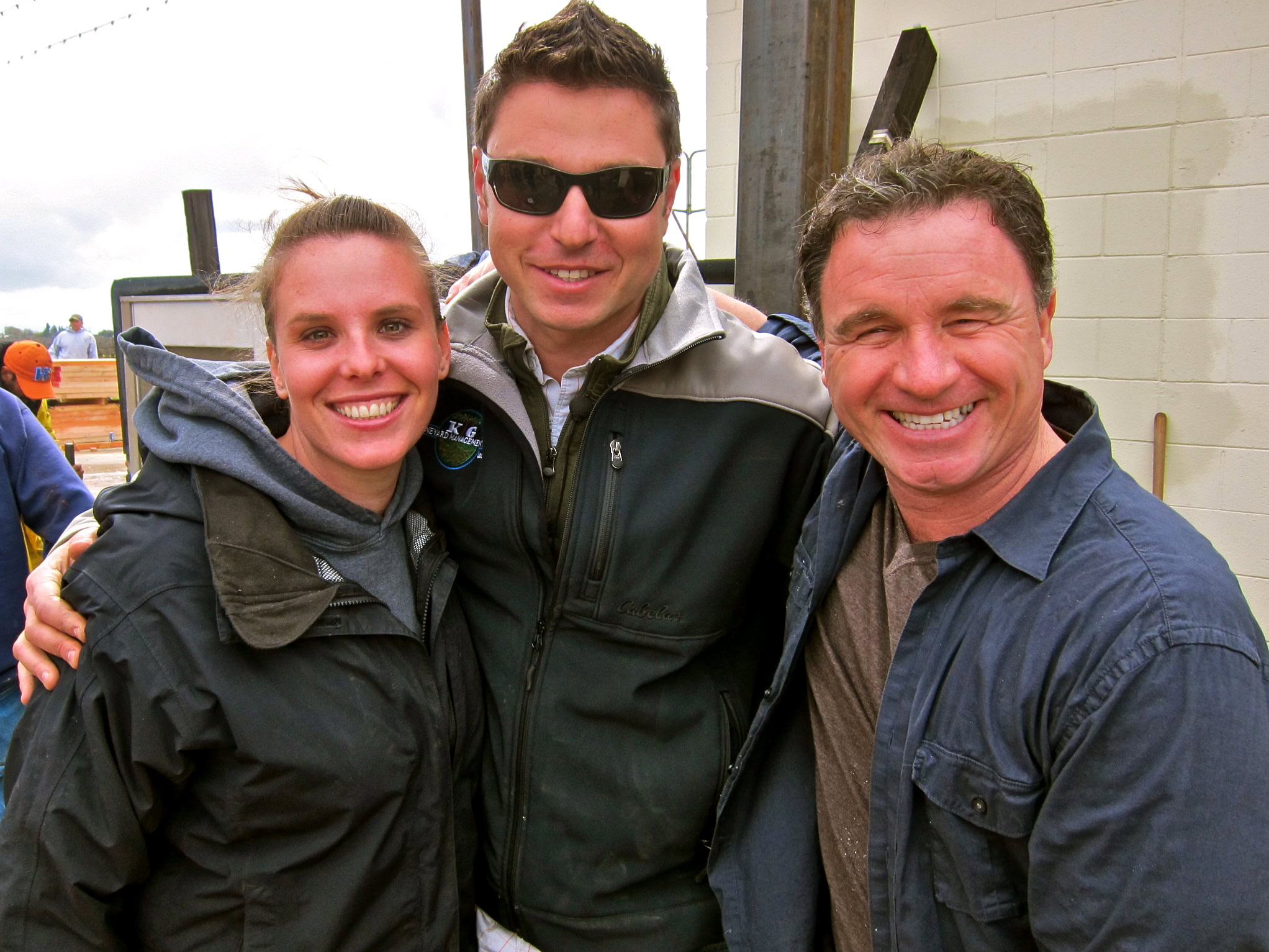 Group of three smiling people at Lodi Winegrape Commission event, outdoors, casual attire, cloudy sky.