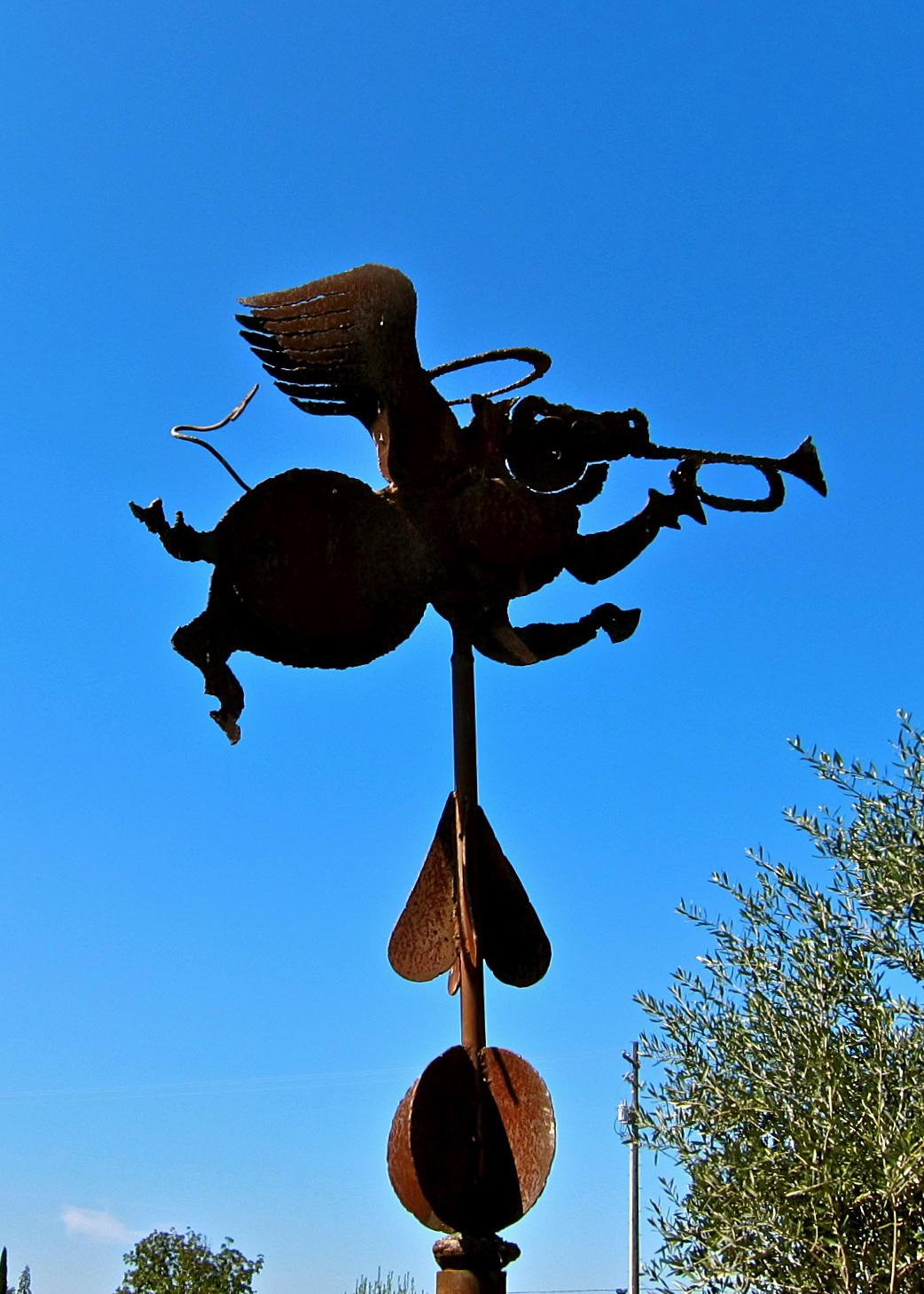 Rusty metal weather vane featuring a flying bird holding a wine glass, set against a clear blue sky.