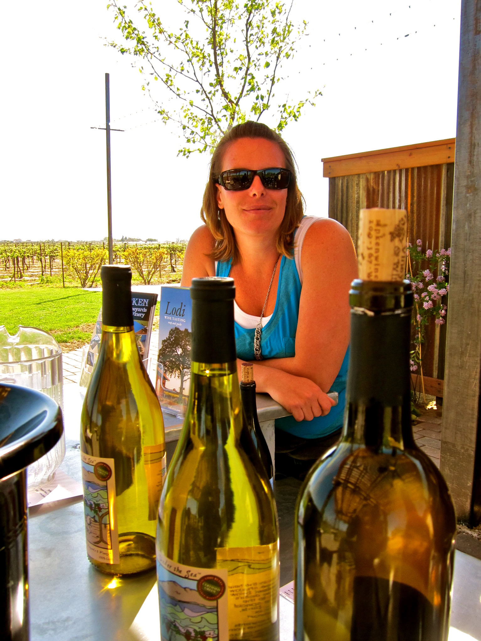 Woman with sunglasses at Lodi wine tasting table with wine bottles and vineyard background.