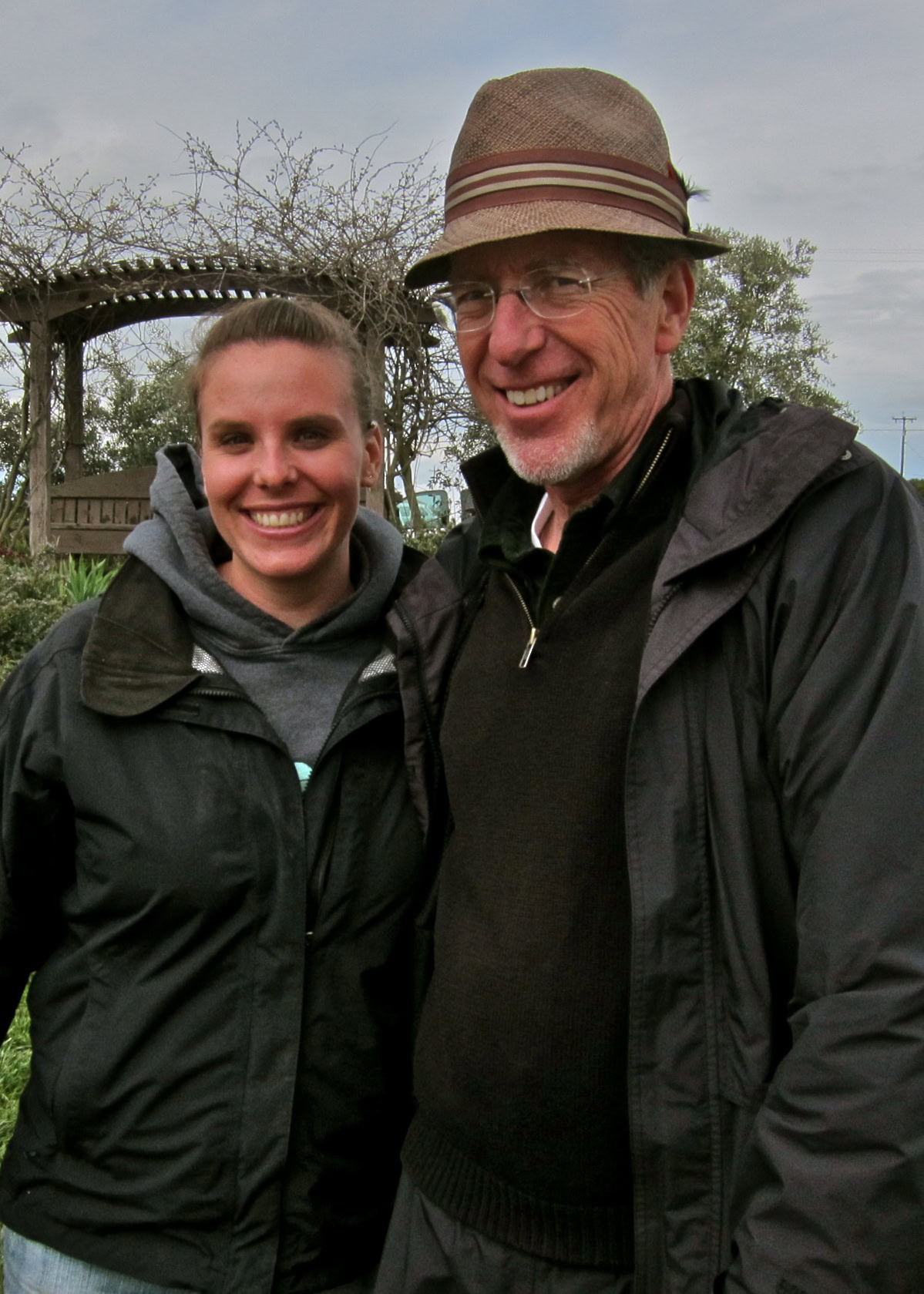 Smiling man and woman outdoors at Lodi Winegrape Commission event, wearing jackets and hats, overcast sky background.