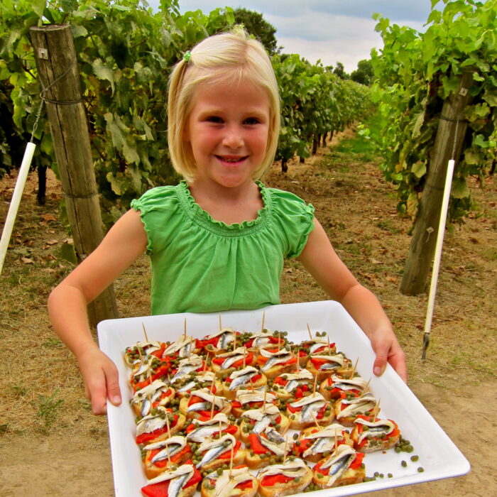 Young girl in a green shirt holding a tray of wine grape snacks in a vineyard setting.