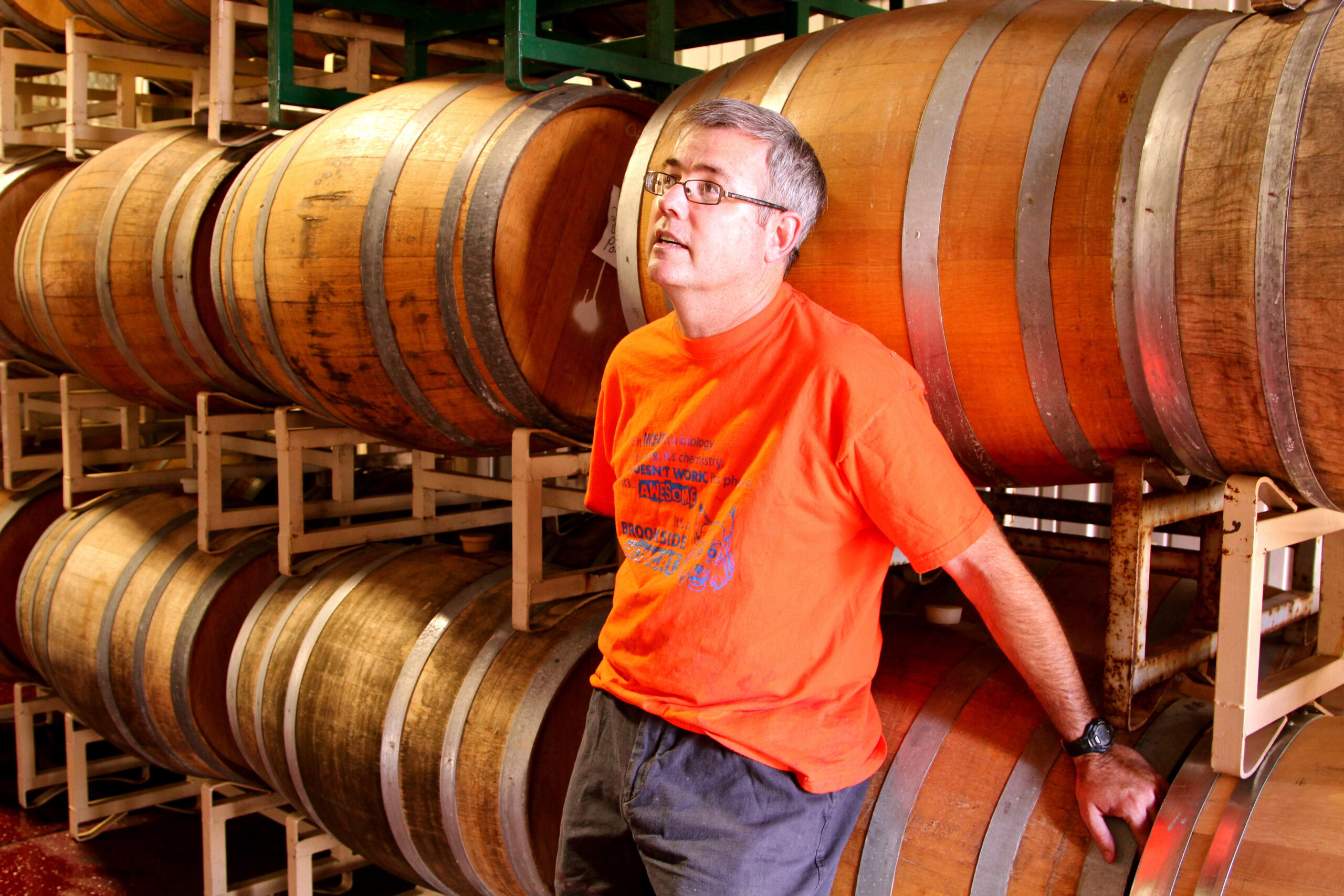 Man in orange shirt at winery with stacked wine barrels in Lodi, California.