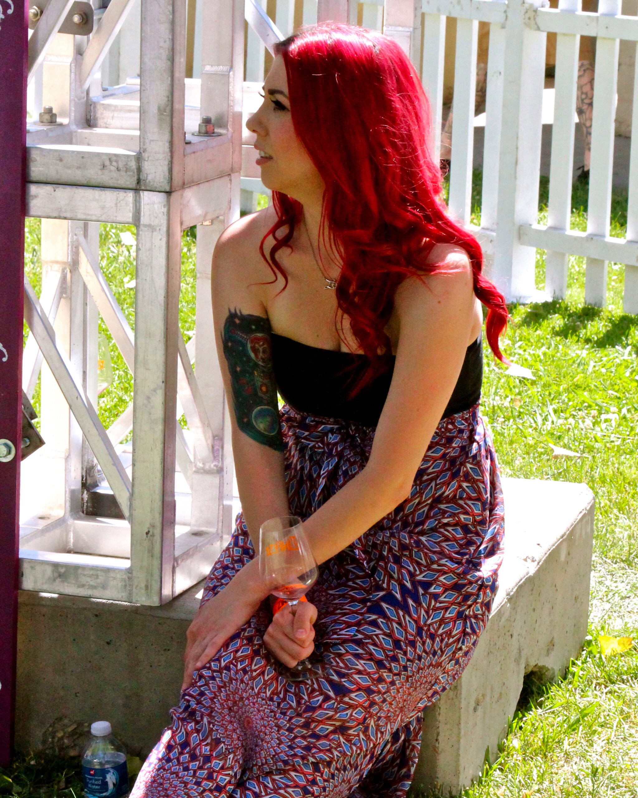 Woman with vibrant red hair holding a glass of rosé wine outdoors in Lodi, California, with vineyard scenery in the backdrop.