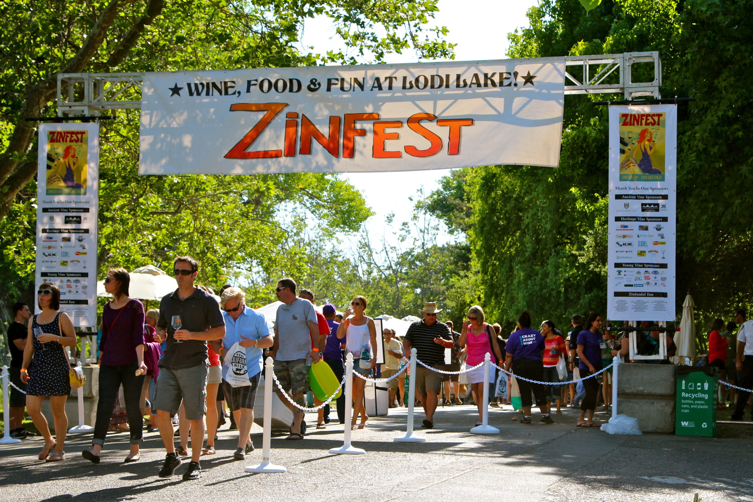 Crowd attending Zinfest wine festival at Lodi Lake with trees and festival banner overhead.