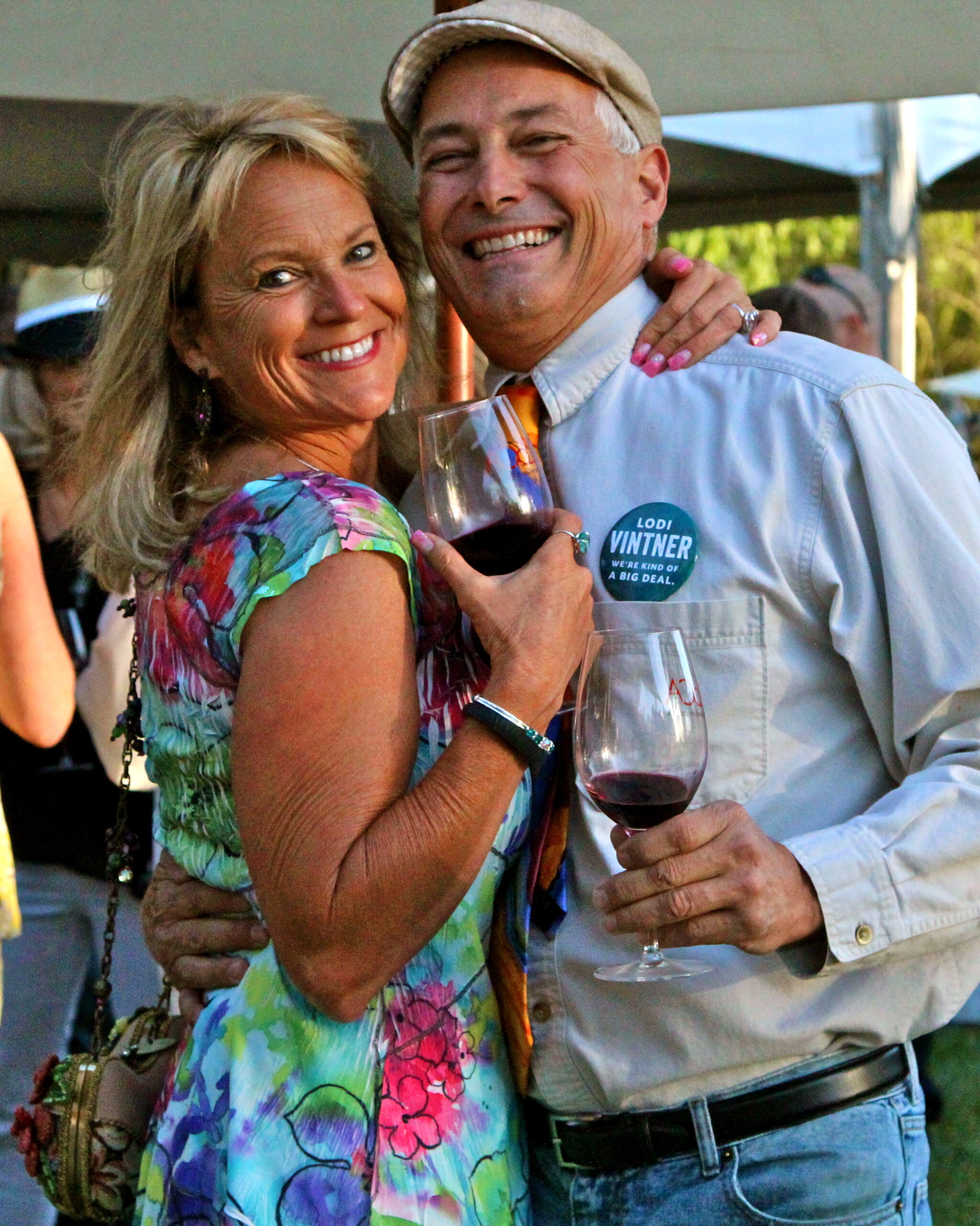 Smiling couple enjoying wine at Lodi Winegrape Commission event, holding glasses of red wine, outdoors setting.