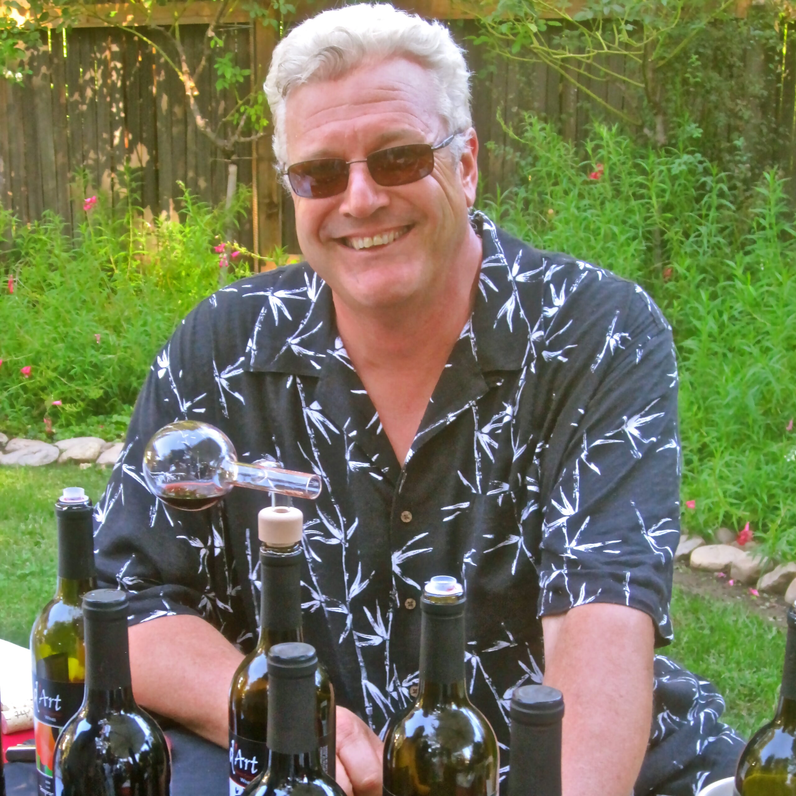 Smiling man in sunglasses at a wine event with bottles and a glass of wine.