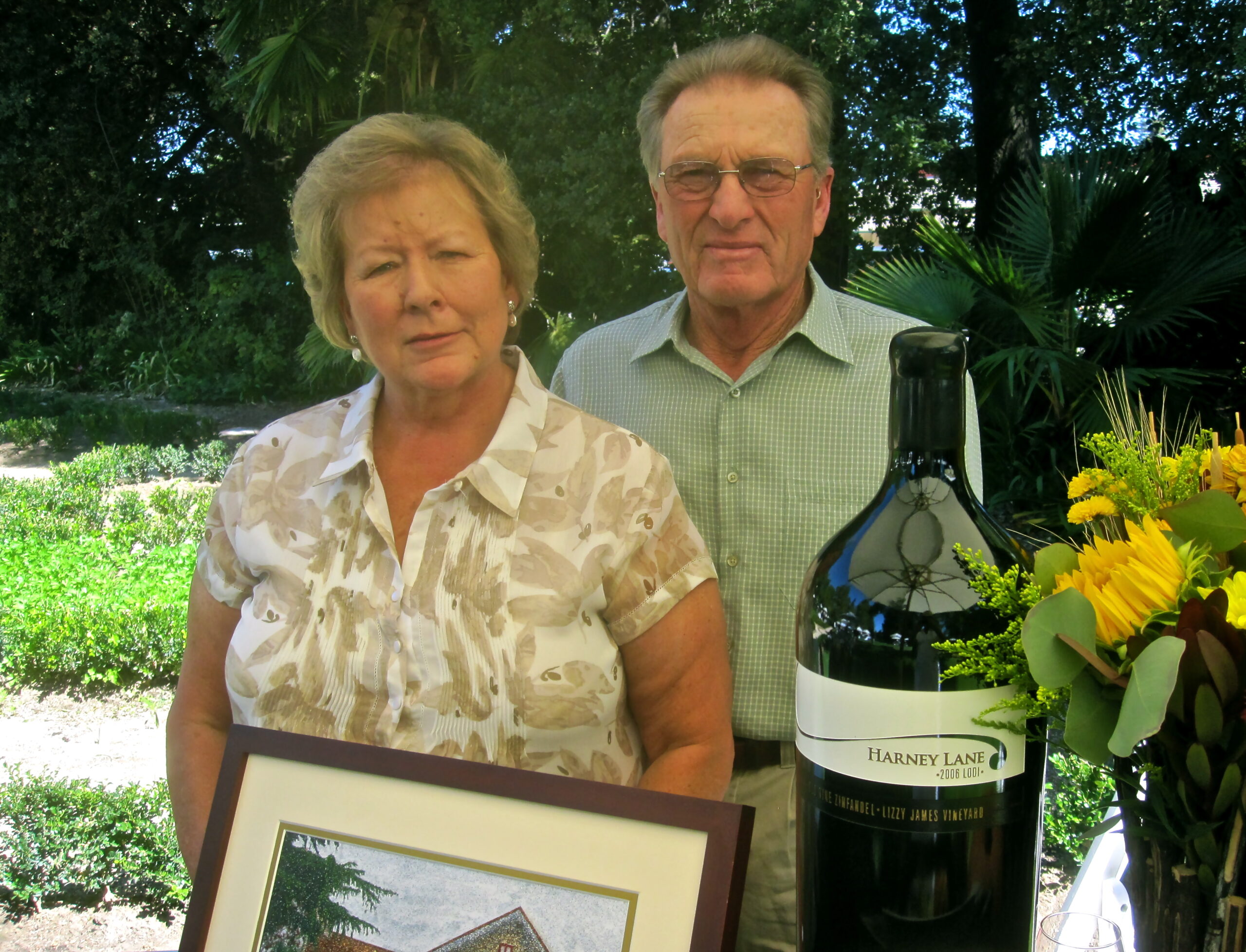 Two elderly individuals standing outdoors with a large wine bottle and a framed photo, surrounded by greenery and flowers.