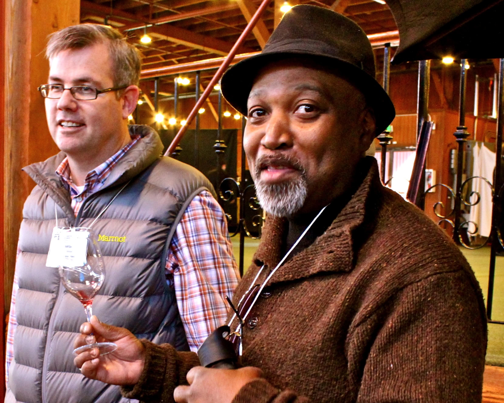 Two men at a wine tasting event, one holding a wine glass, in a winery setting with warm lighting.
