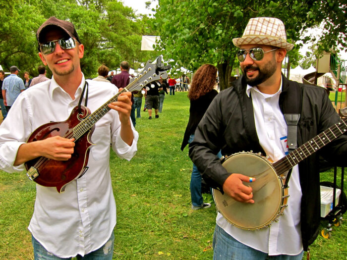 Two musicians playing string instruments at a Lodi wine event with trees and people in the background.