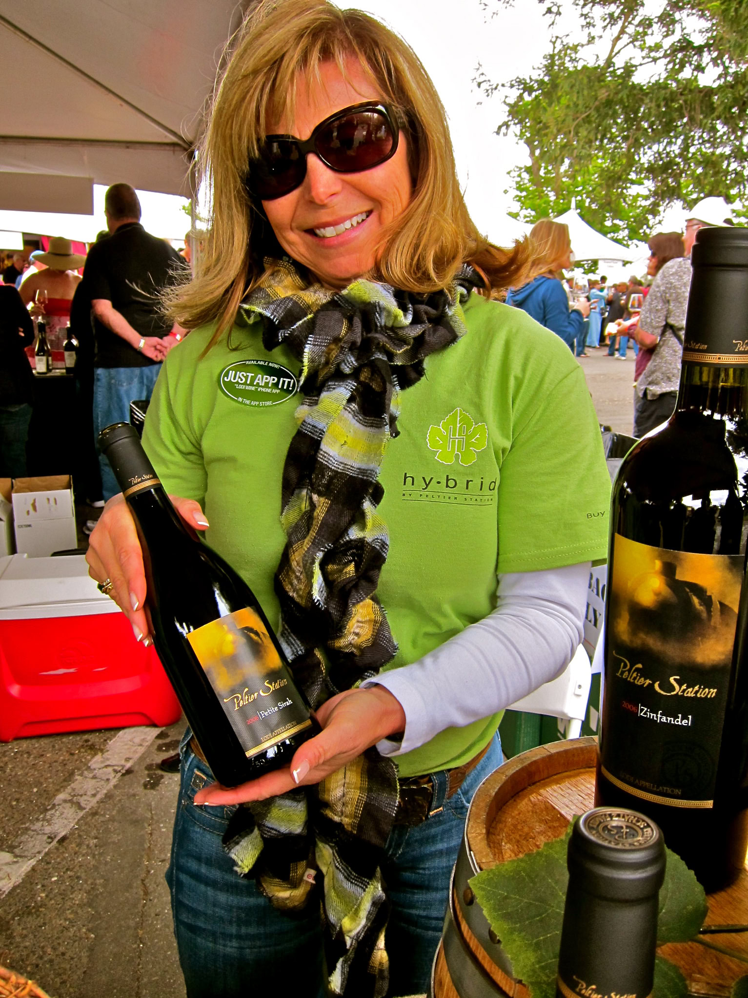 Woman holding a bottle of wine at a Lodi wine event, smiling and wearing sunglasses and a green shirt.