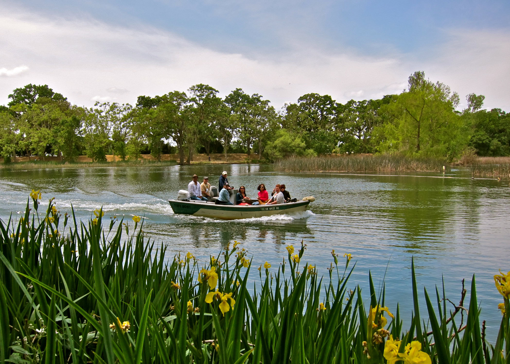 People enjoying a boat ride on a river surrounded by lush greenery and yellow flowers in Lodi, California.