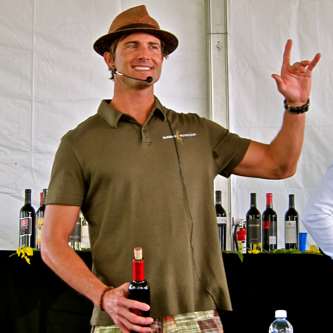 Man in a brown hat and green polo speaking at a wine event with bottles in the background.