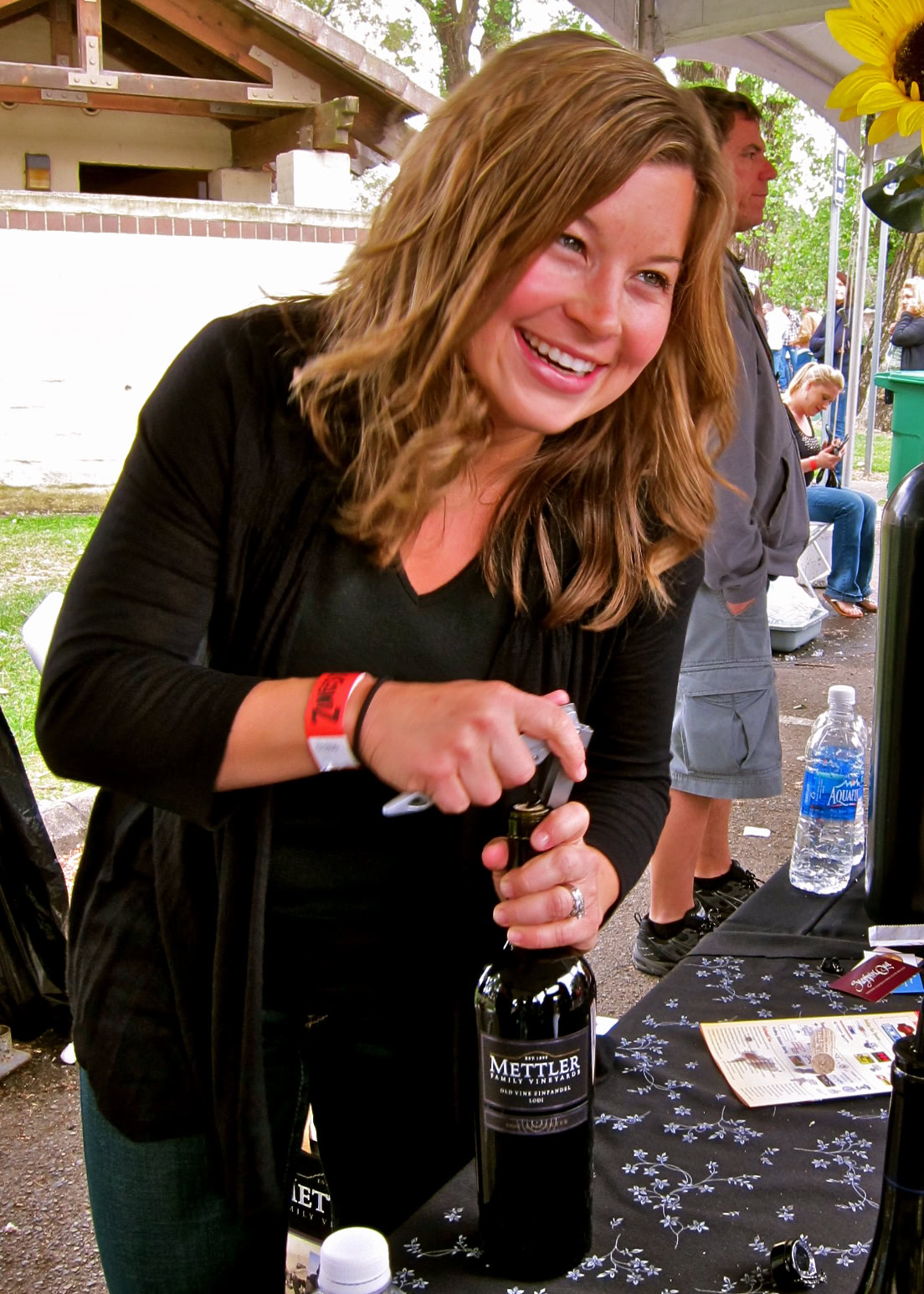 Smiling woman opening a wine bottle at a Lodi wine event, with people and outdoor setting in background.
