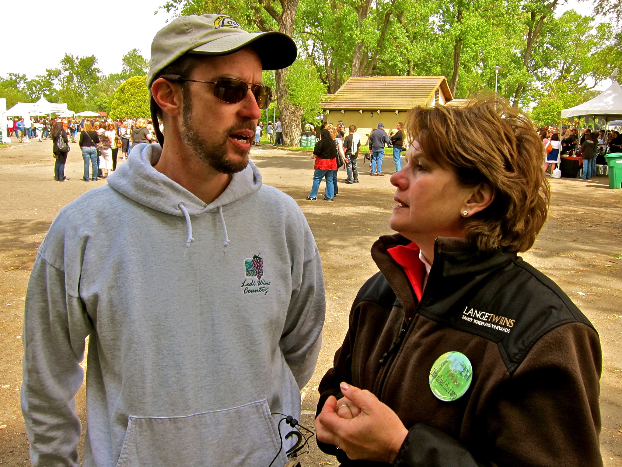 Man and woman talking at Lodi Winegrape Commission outdoor event with trees and tents in background.