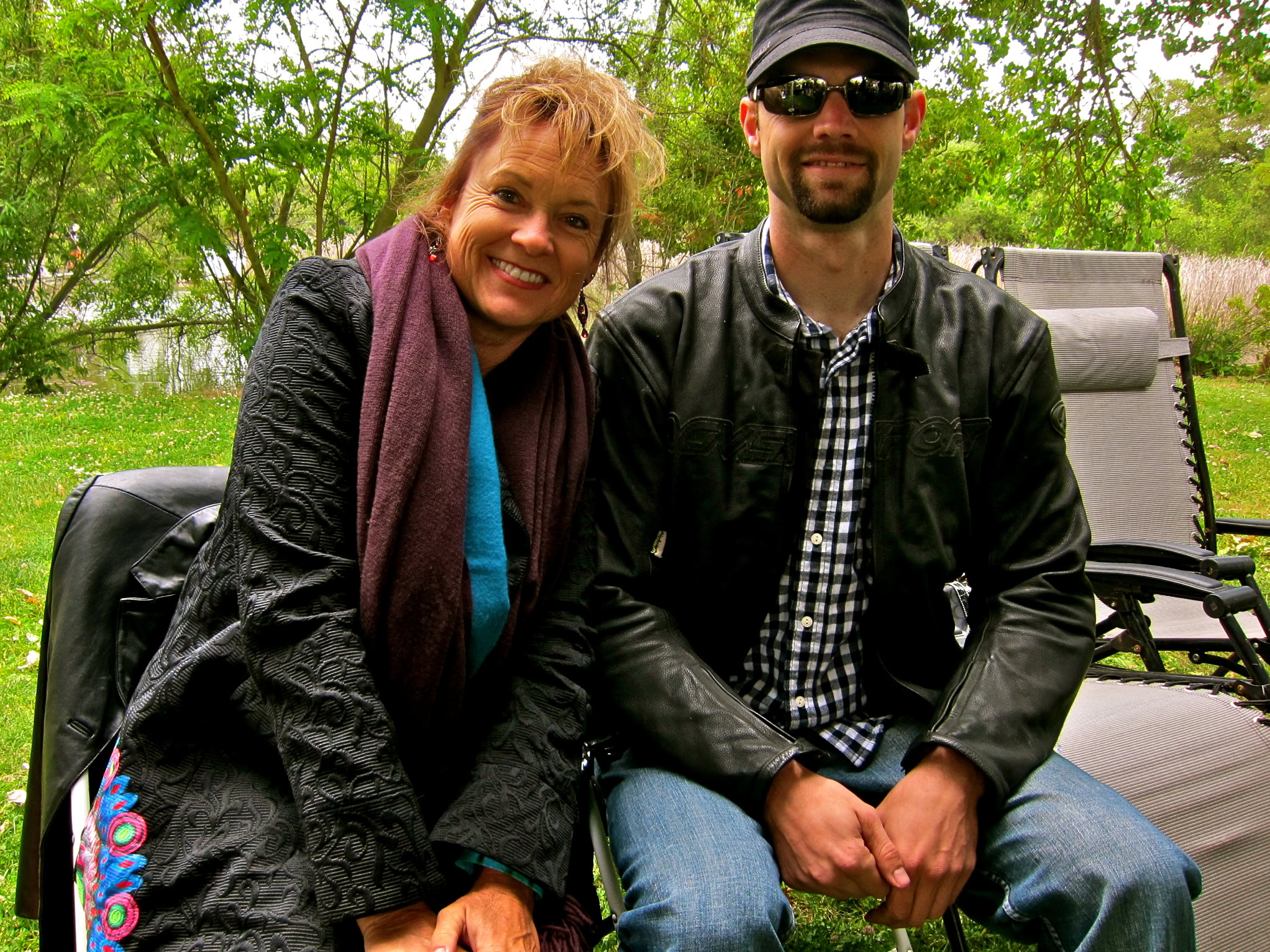 Two people sitting outdoors near a lake, smiling, with trees in the background, representing Lodi Winegrape Commission.