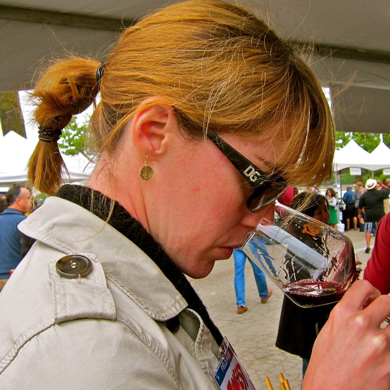 Woman tasting red wine at Lodi Winegrape Commission event, outdoor setting with crowd and tents.
