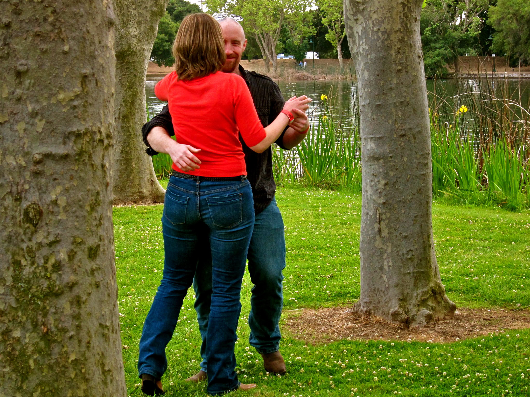 A couple dancing outdoors in a park with trees and a pond in the background.