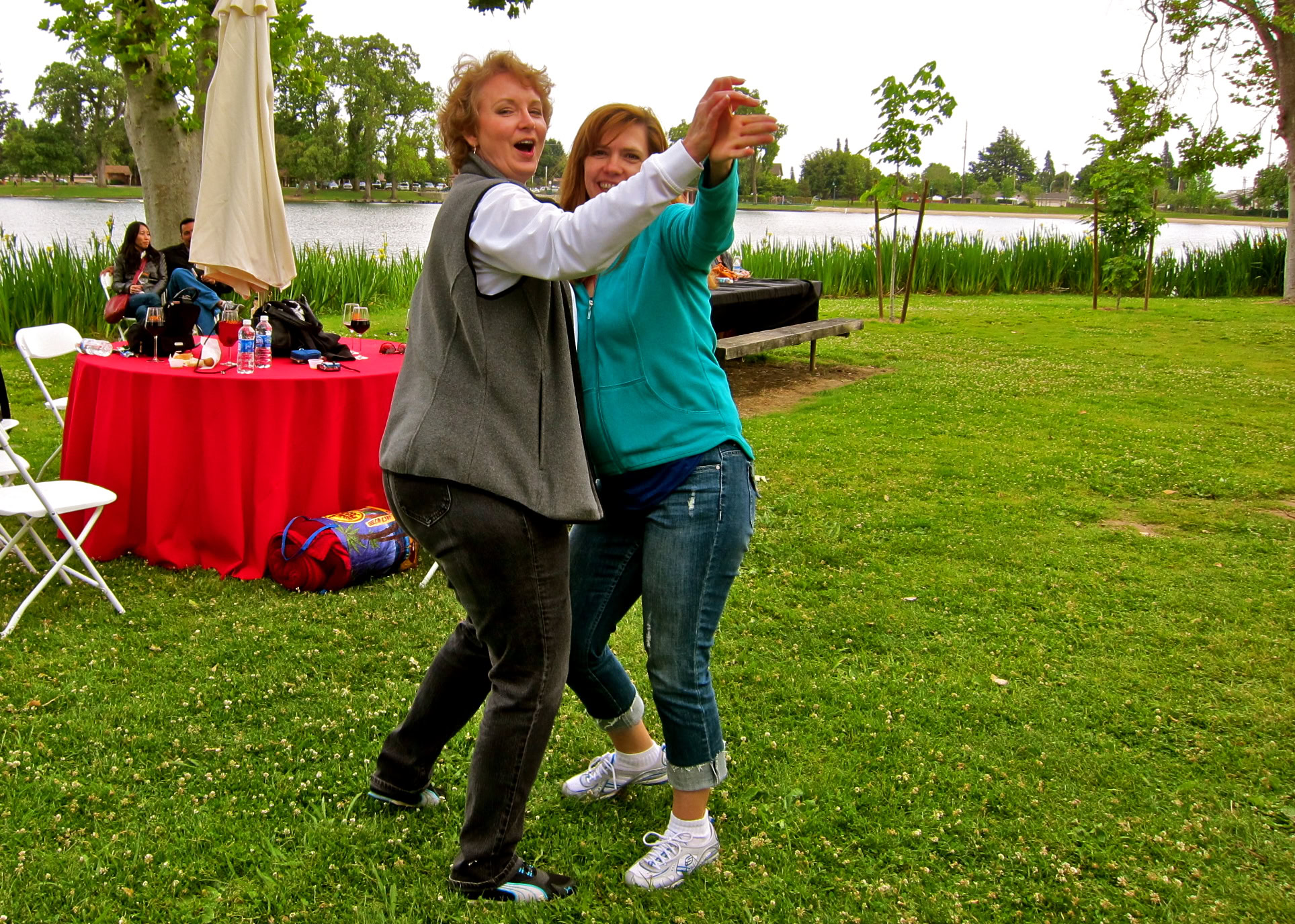 Two women taking a selfie outdoors near a lake with a red table and people in the background.
