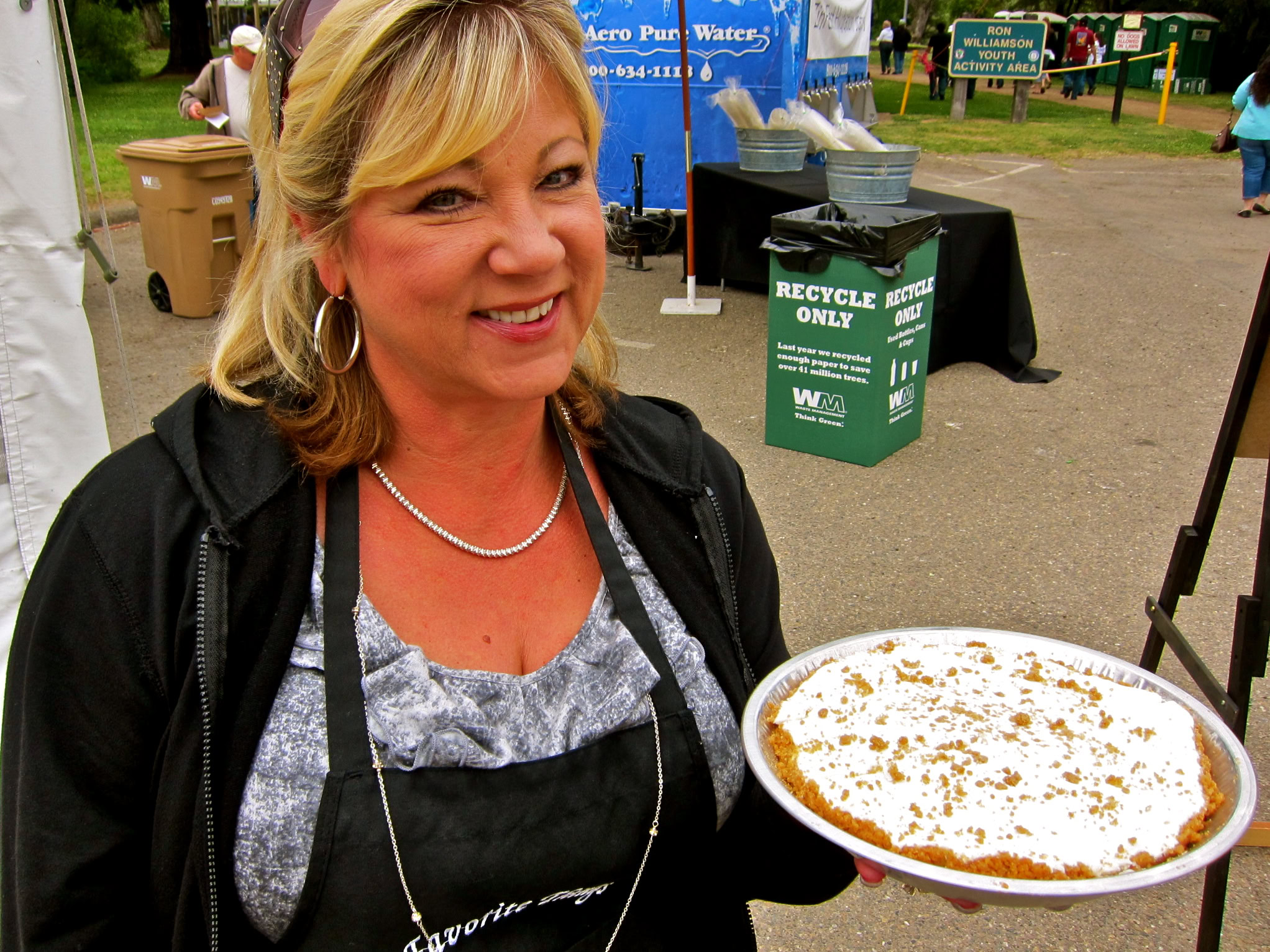 Smiling woman holding a slice of pie at a Lodi wine event, with recycling bins and event tents in the background.