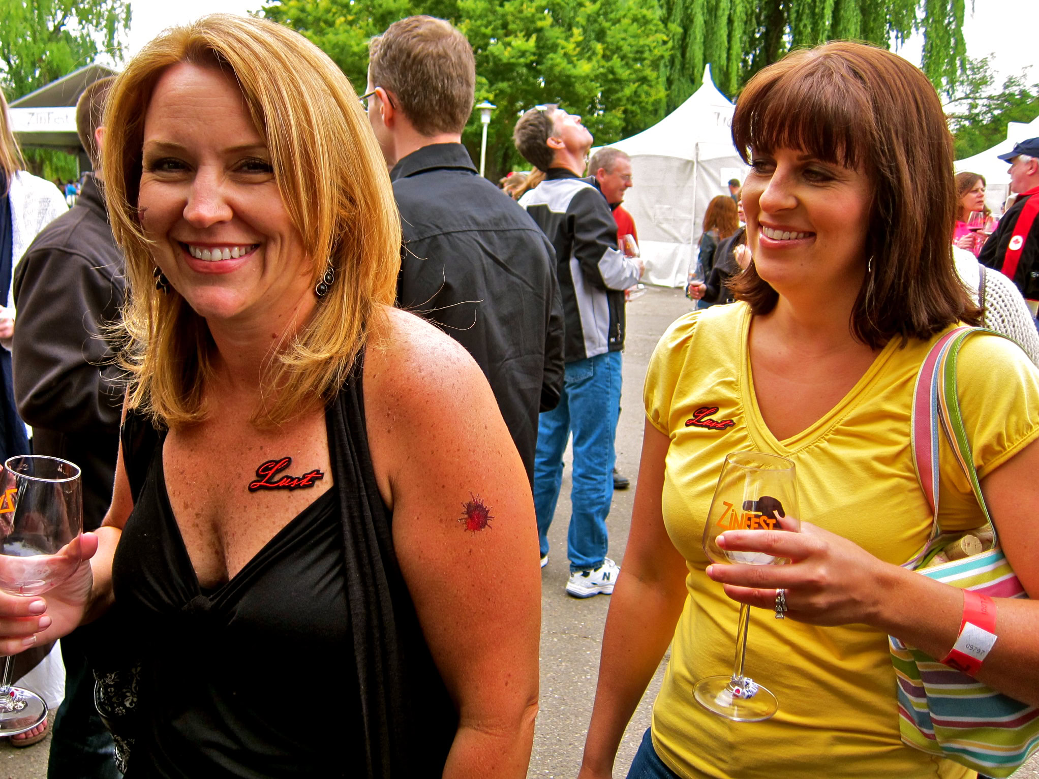 Two women enjoying wine at the Lodi Winegrape Festival outdoors with trees and tents in the background.