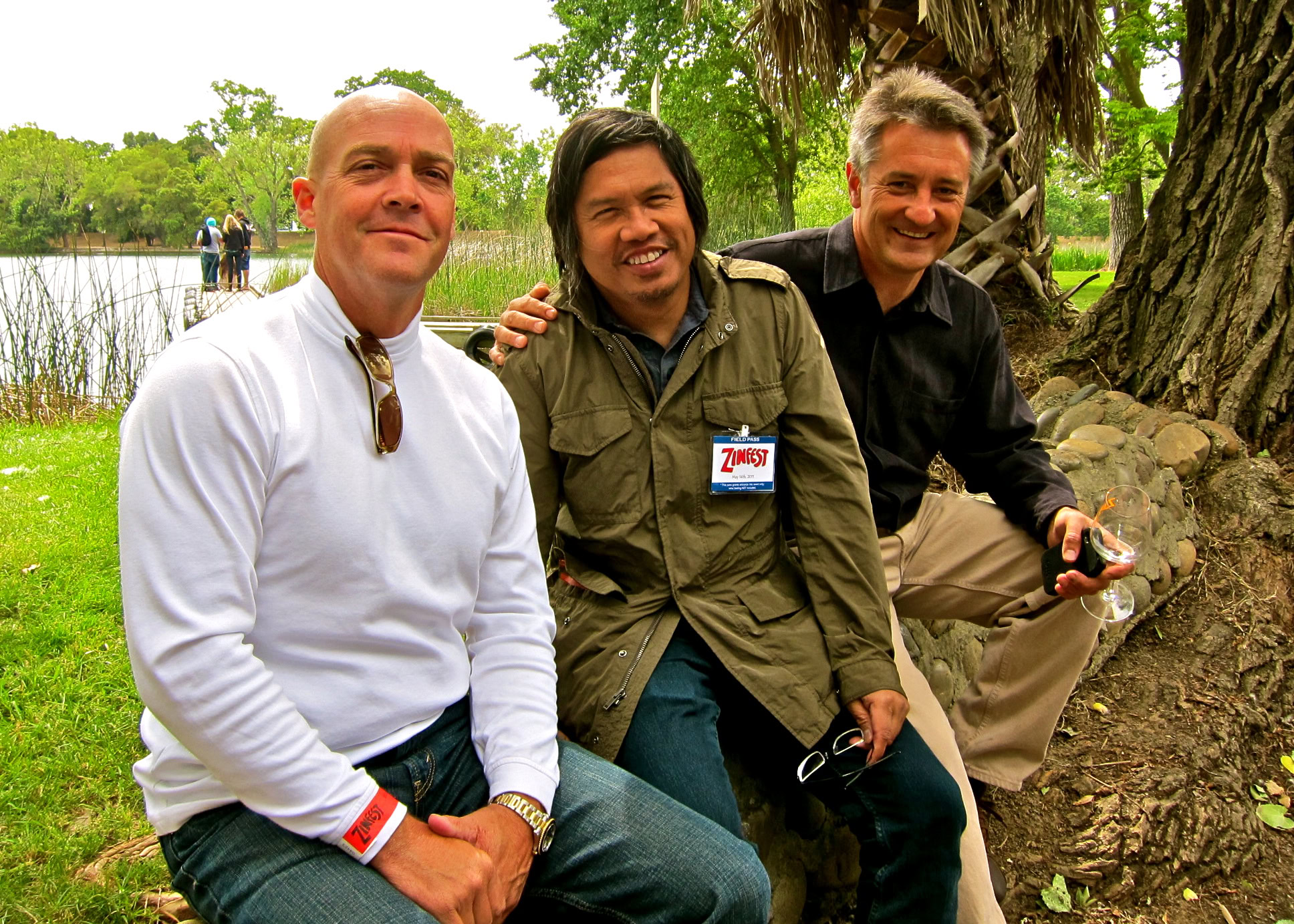 Three men sitting outdoors near a lake, smiling, with trees and water in the background.
