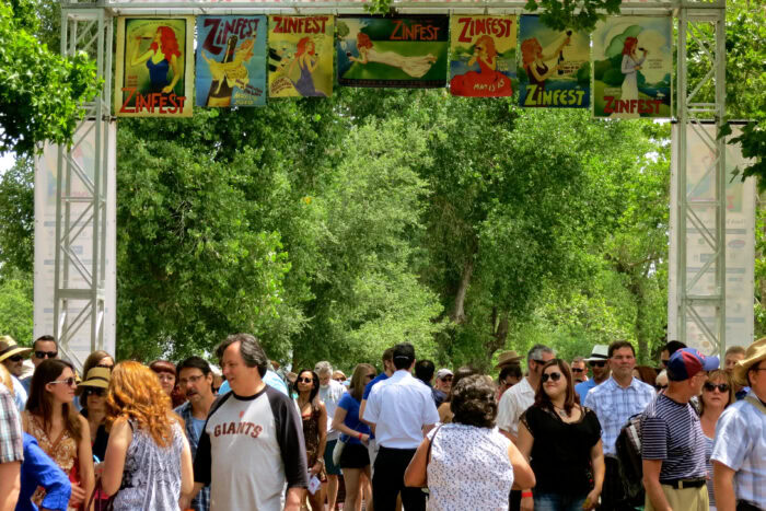 Crowd attending Zinfest festival with banners and lush green trees in the background.