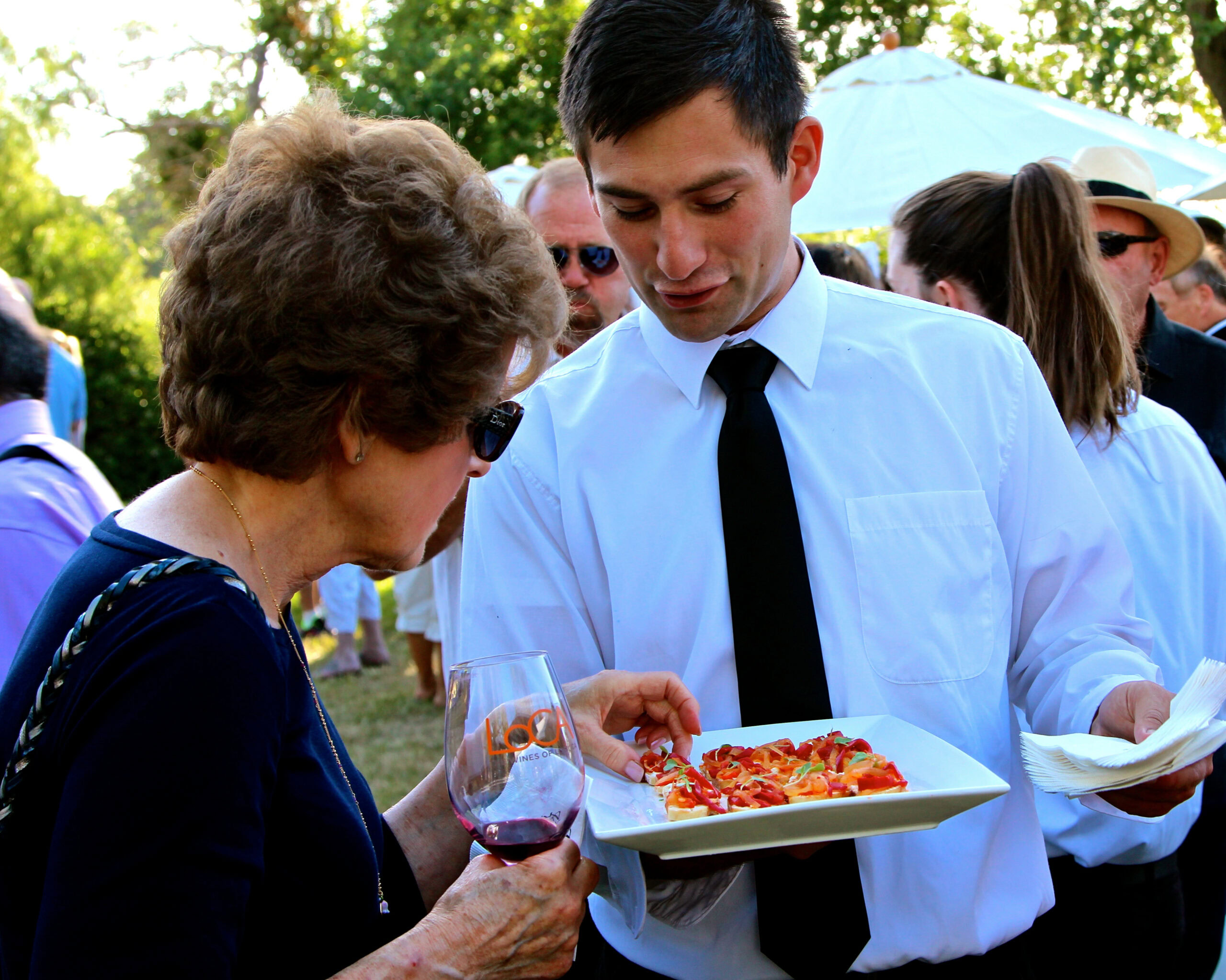 Young man serving pizza at Lodi Winegrape Commission outdoor event with attendees in background.