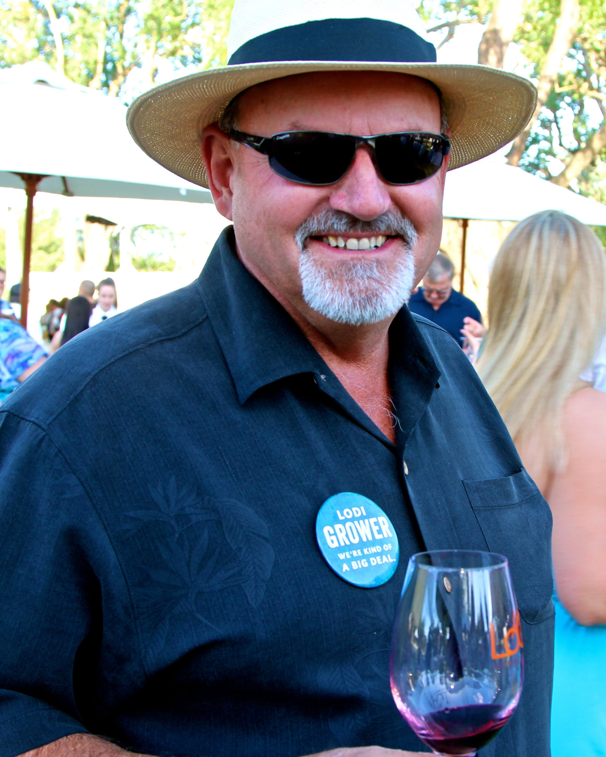 Smiling man with sunglasses and straw hat holding a glass of red wine at outdoor wine event.