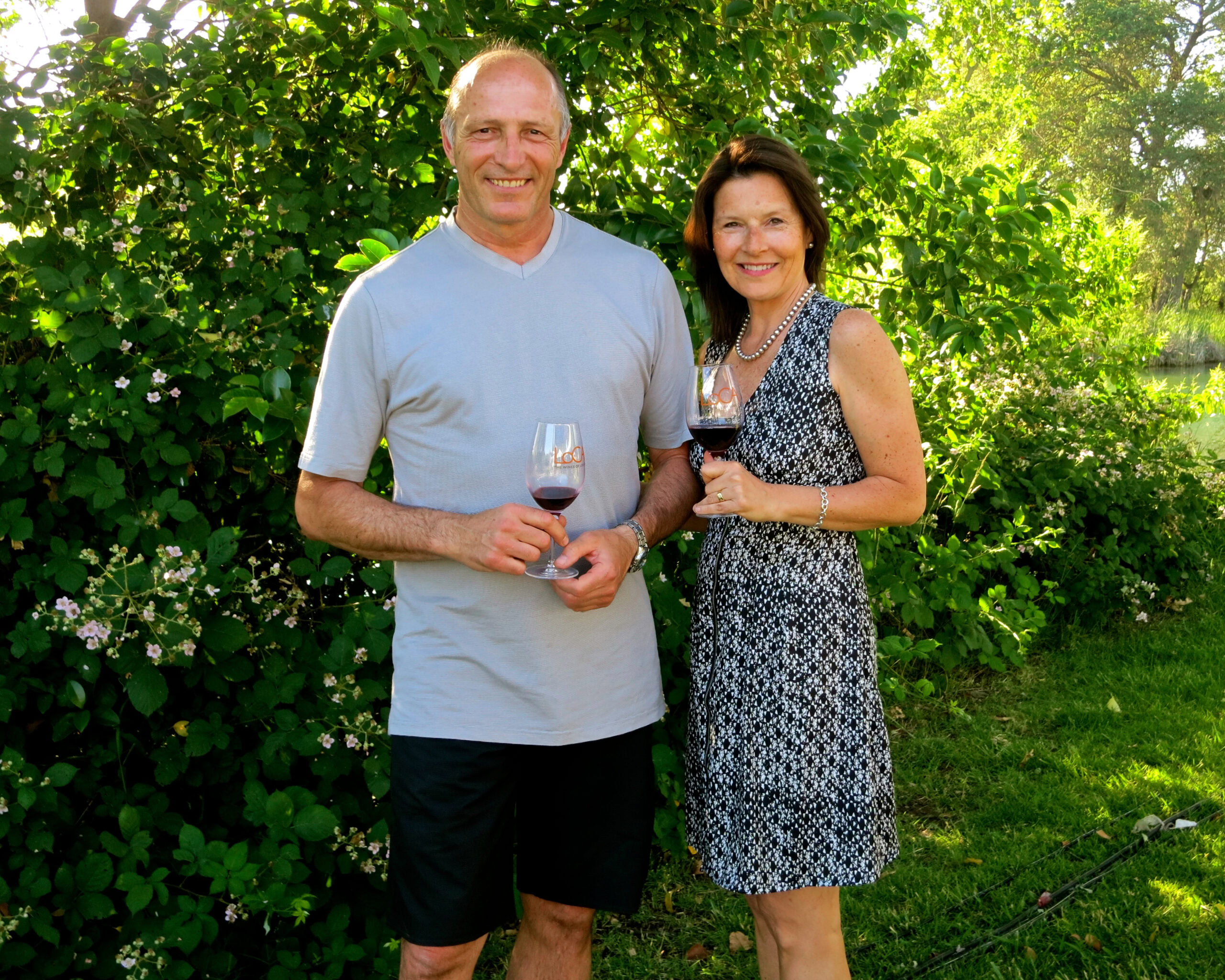 Smiling couple enjoying wine outdoors in lush green vineyard setting.