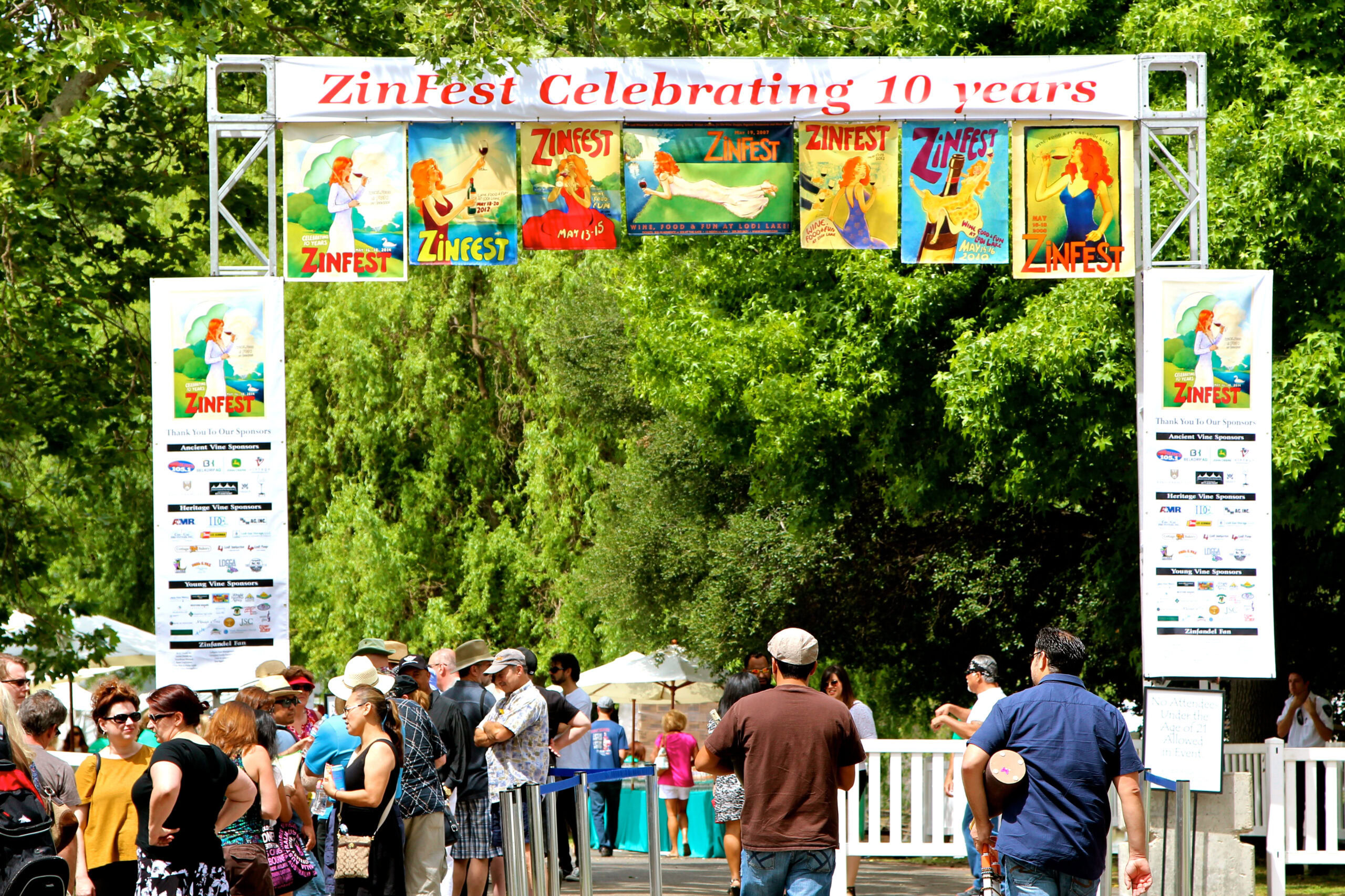 Crowd attending Zinfest wine festival under a decorated archway with banners and trees in the background.