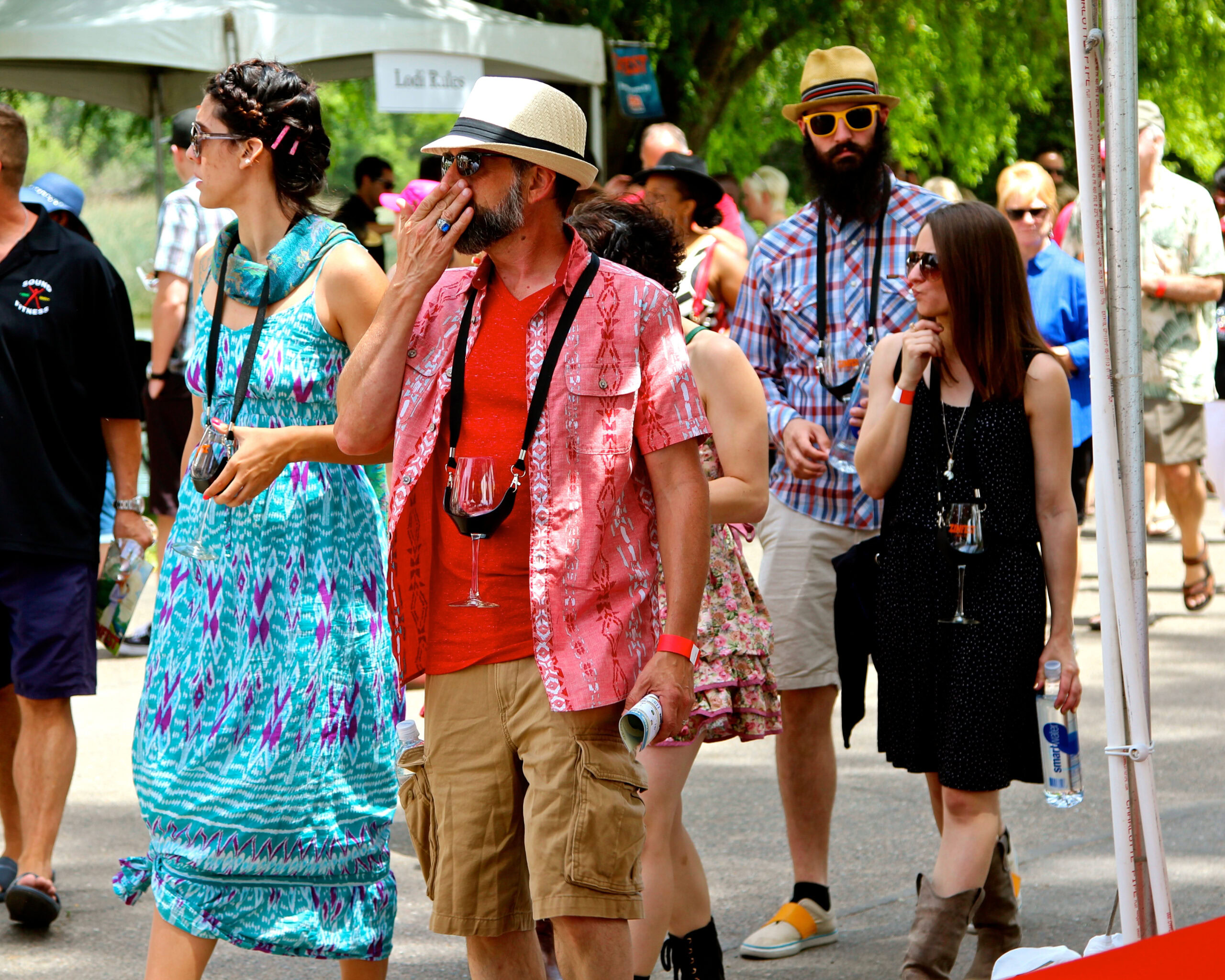 Group of people enjoying a wine event outdoors, wearing summer clothes and sunglasses, with trees and tents in the background.