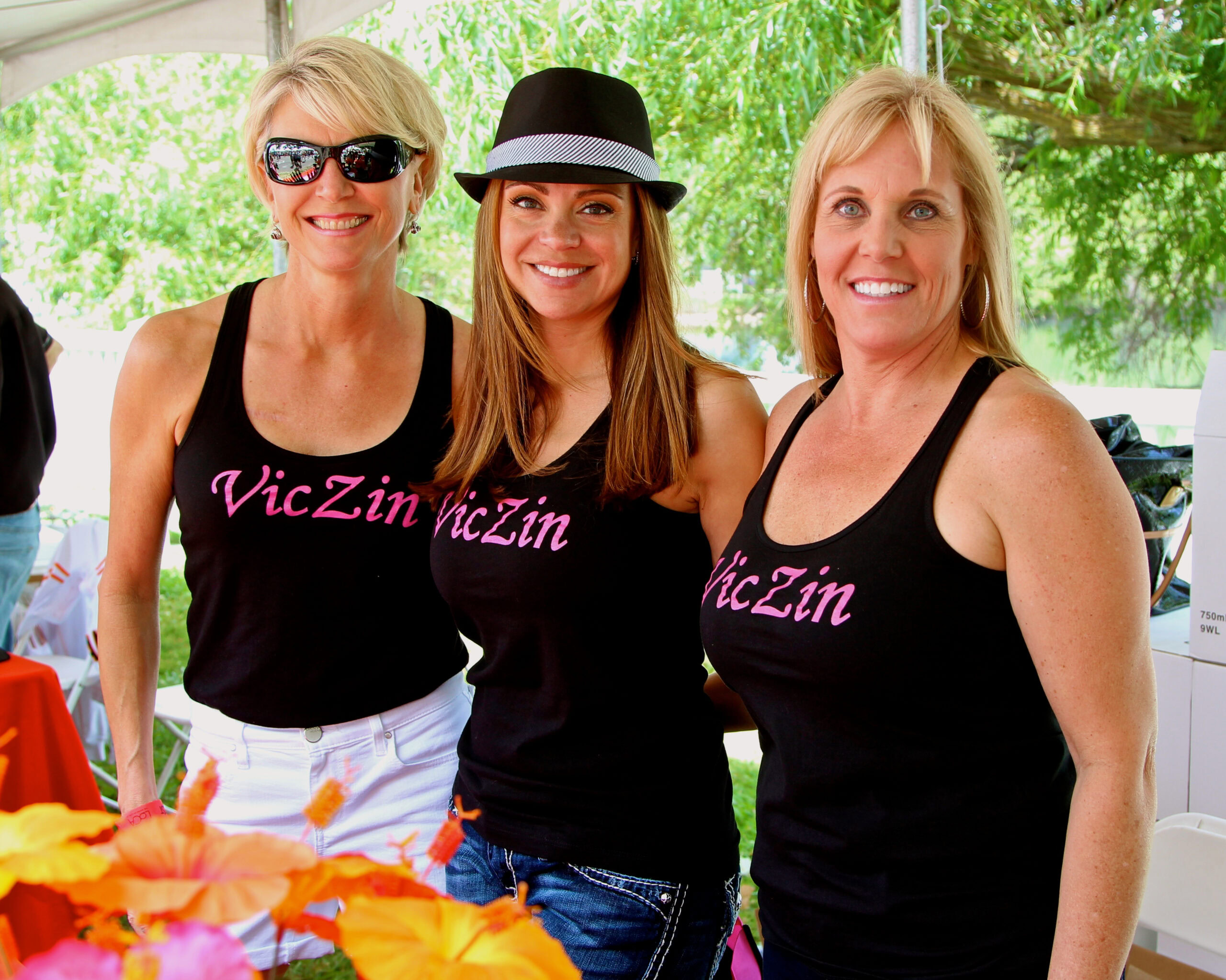 Three women at Lodi Winegrape Commission event wearing black tank tops with pink text, smiling outdoors.