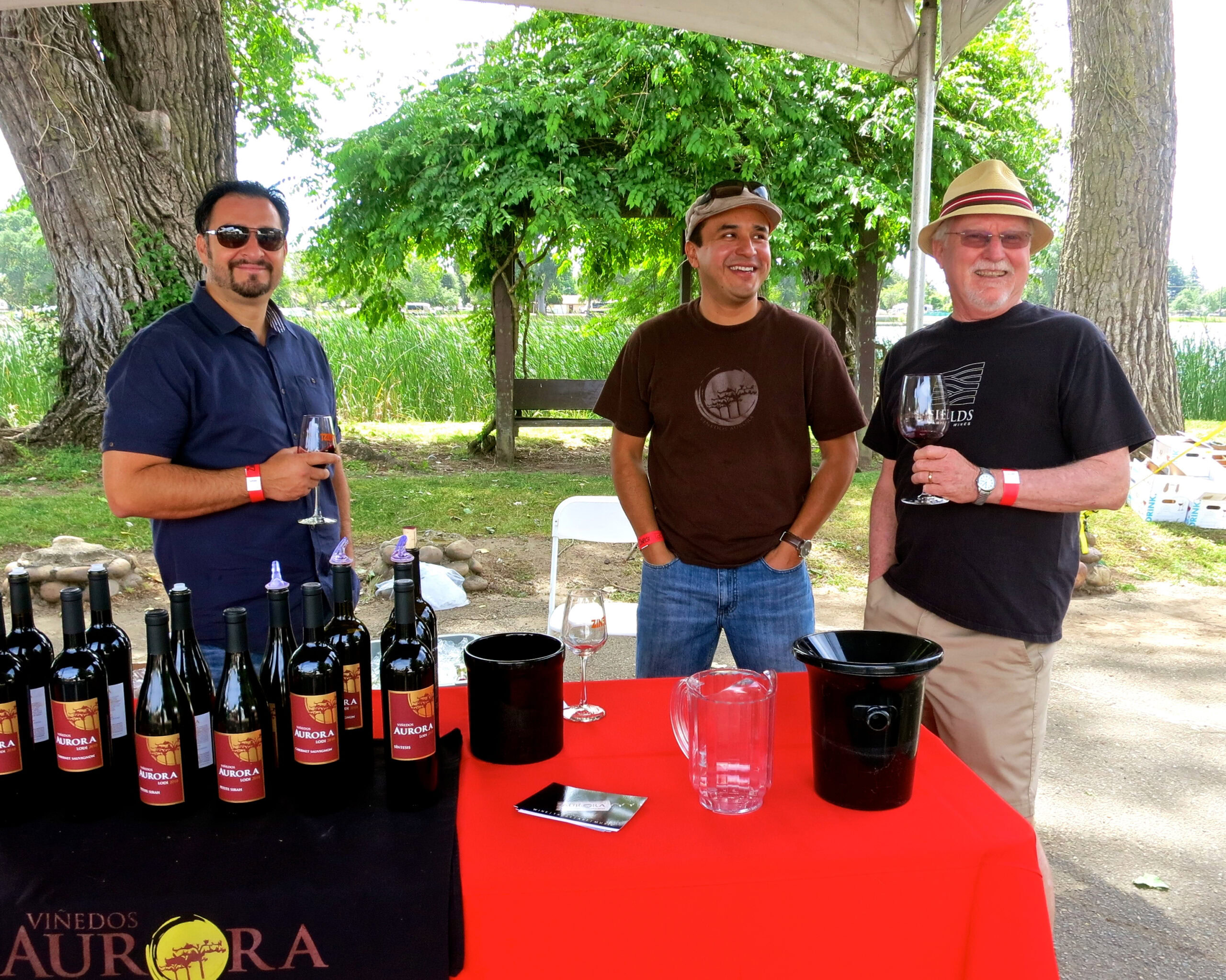Three men at a wine tasting event outdoors with wine bottles on a table under a canopy.