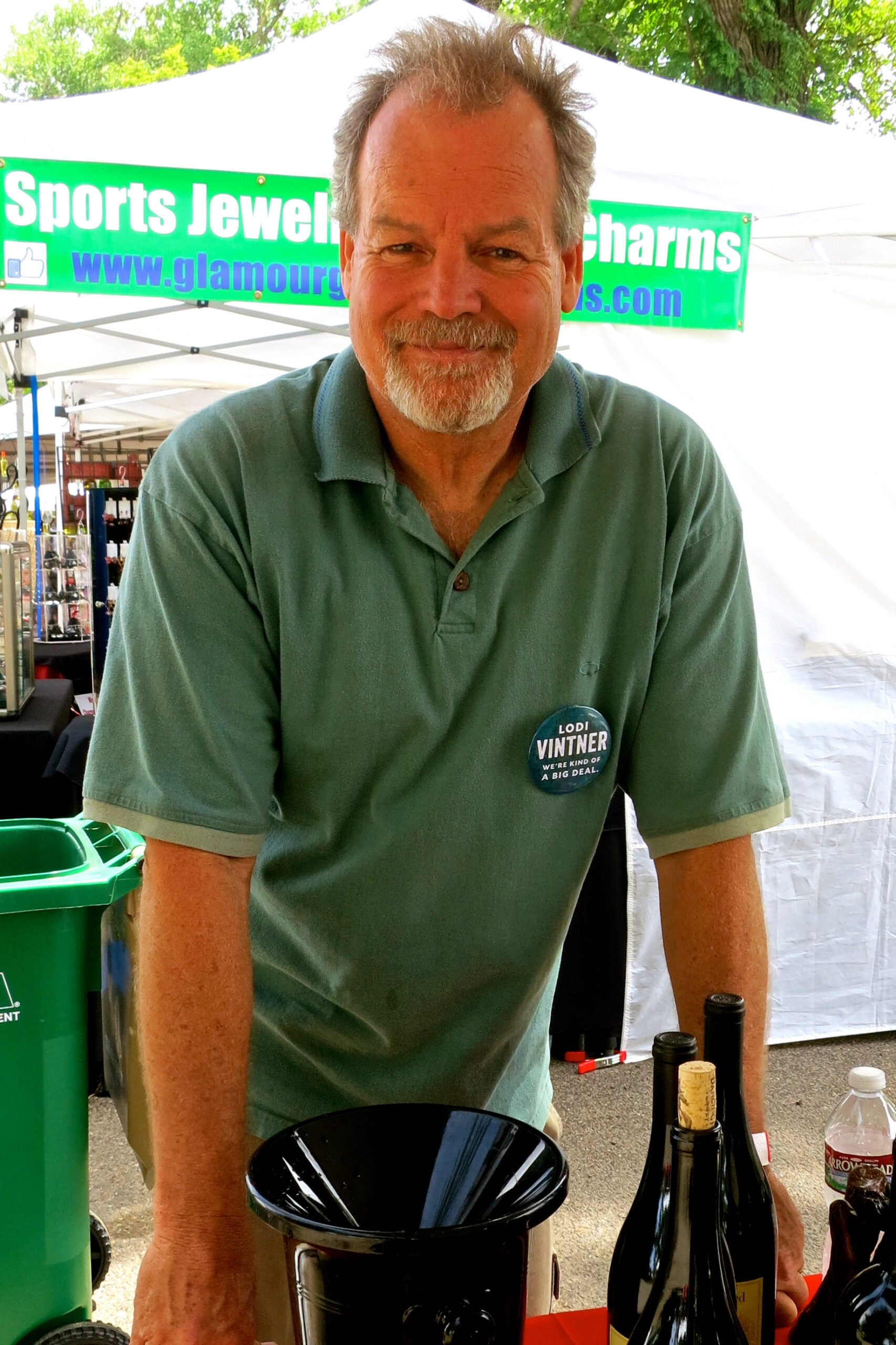 Smiling man at Lodi Winegrape Commission event with wine bottles and booth in background.