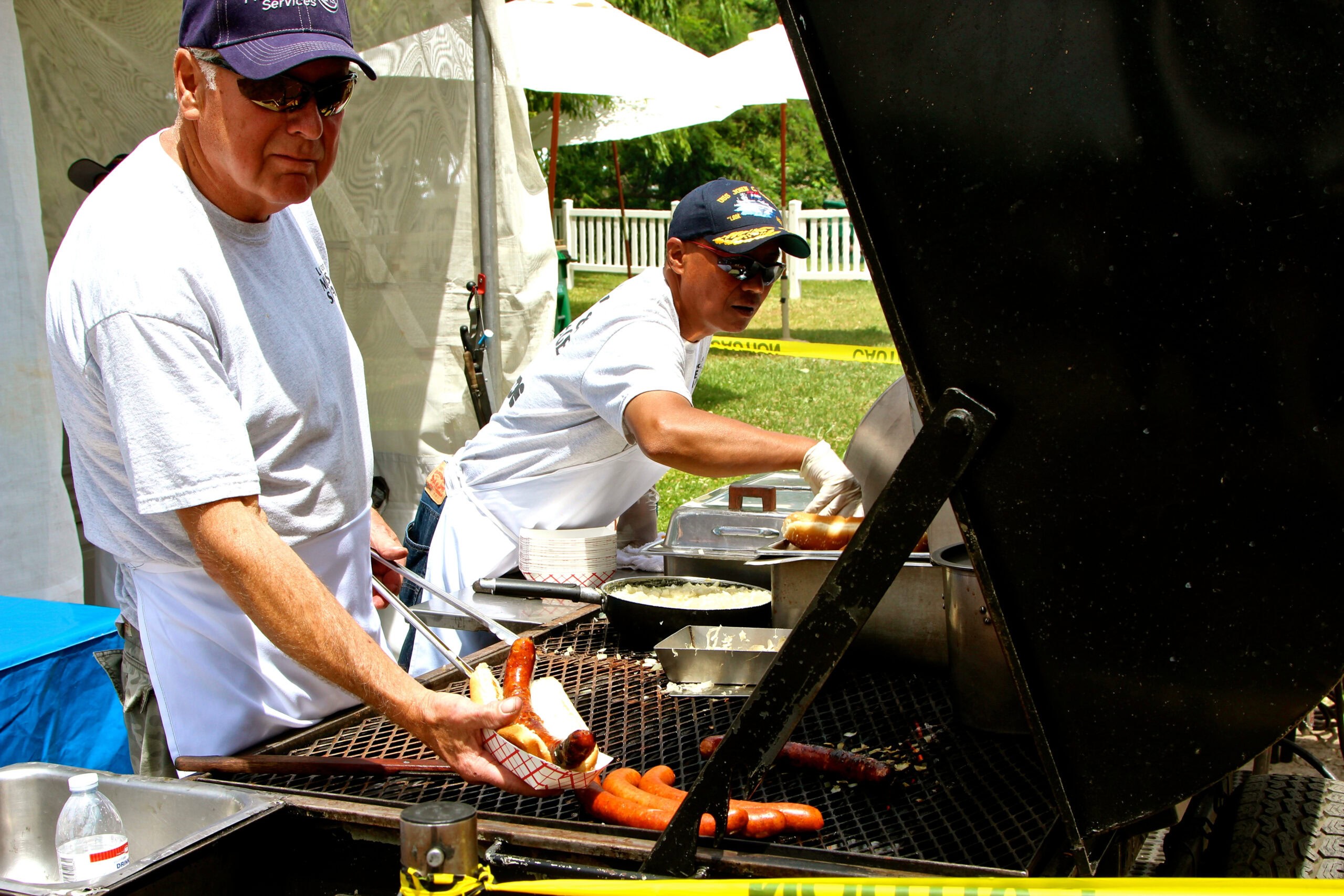 Two men grilling sausages and vegetables at a Lodi Winegrape Commission event outdoors.