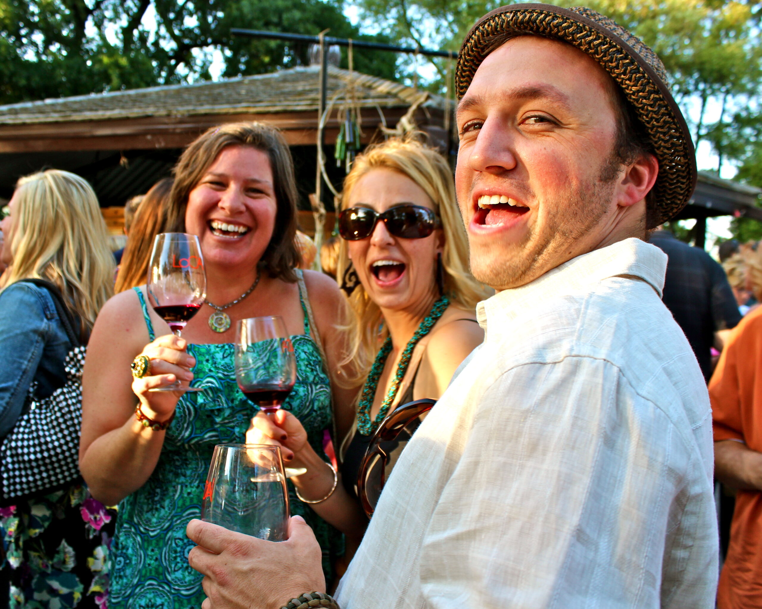 Group of people enjoying wine at an outdoor event, smiling and holding wine glasses, with trees and a rustic structure in the.