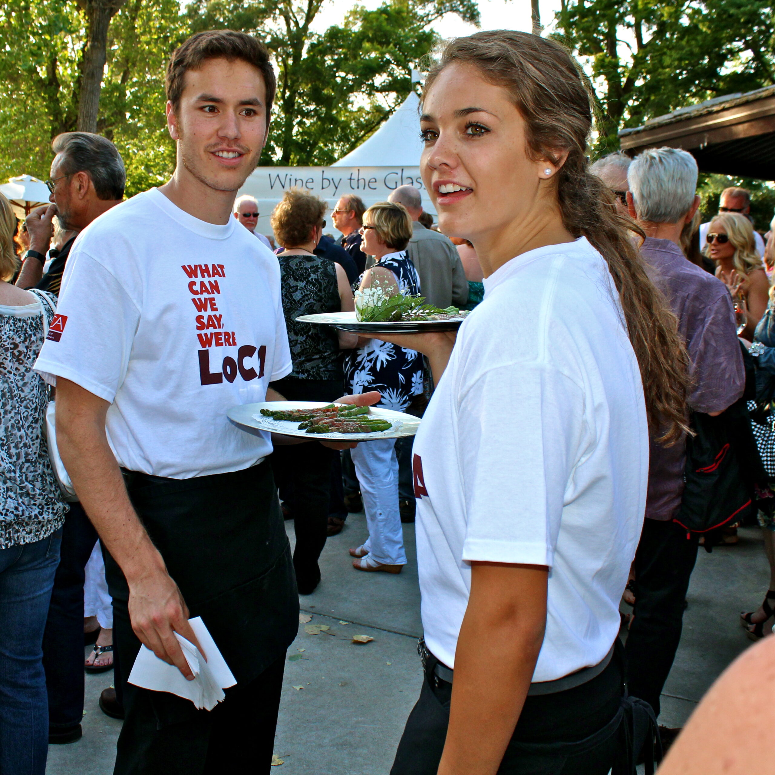 Two young servers at a Lodi wine event holding plates of food, with a crowd and trees in the background.