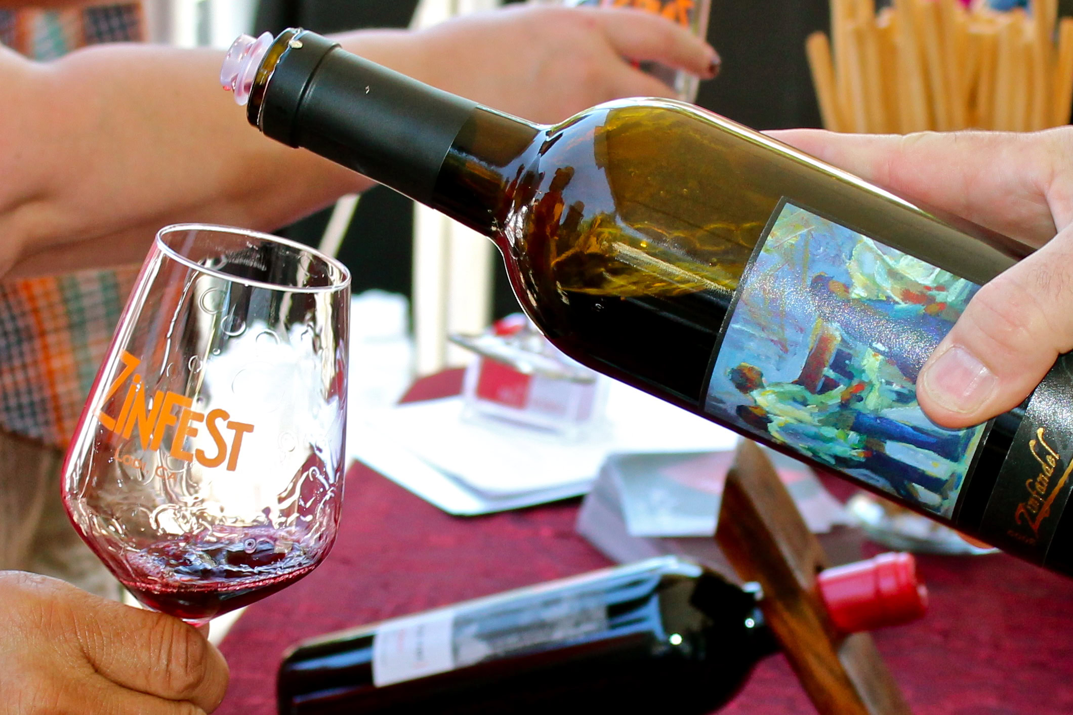 Person pouring red wine into a glass at a wine tasting event, with wine bottles and a table in the background.