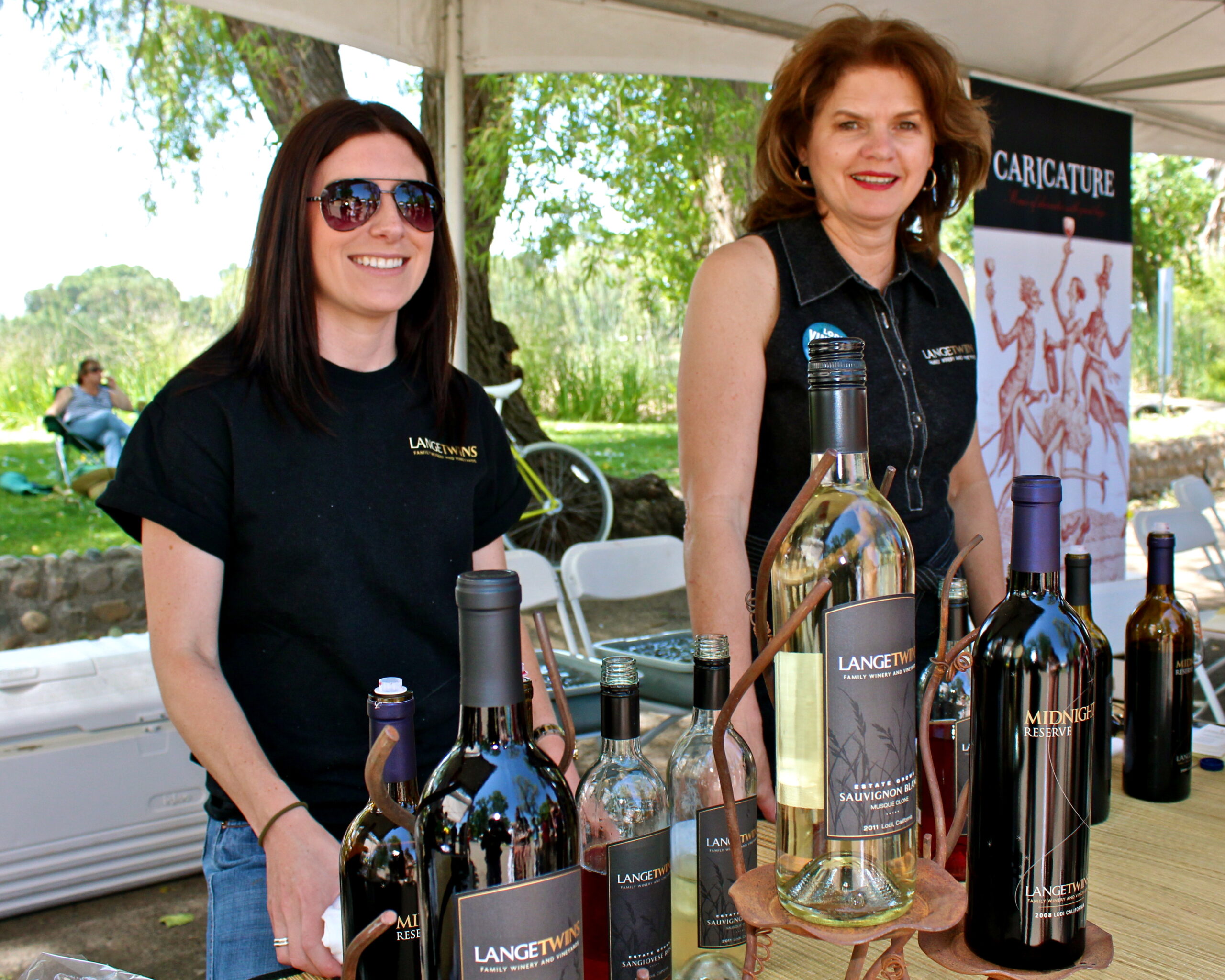 Two women at a wine tasting booth with bottles of wine and a caricature artist banner in the background.