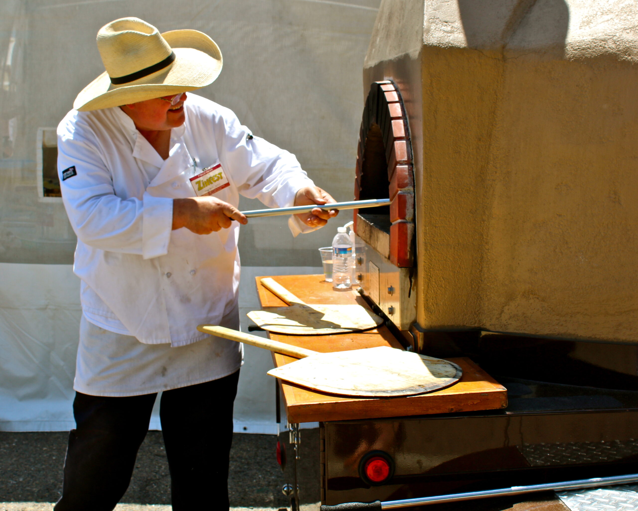 Person baking pizza in a wood-fired oven at a Lodi wine event, wearing a large sun hat and white chef coat.