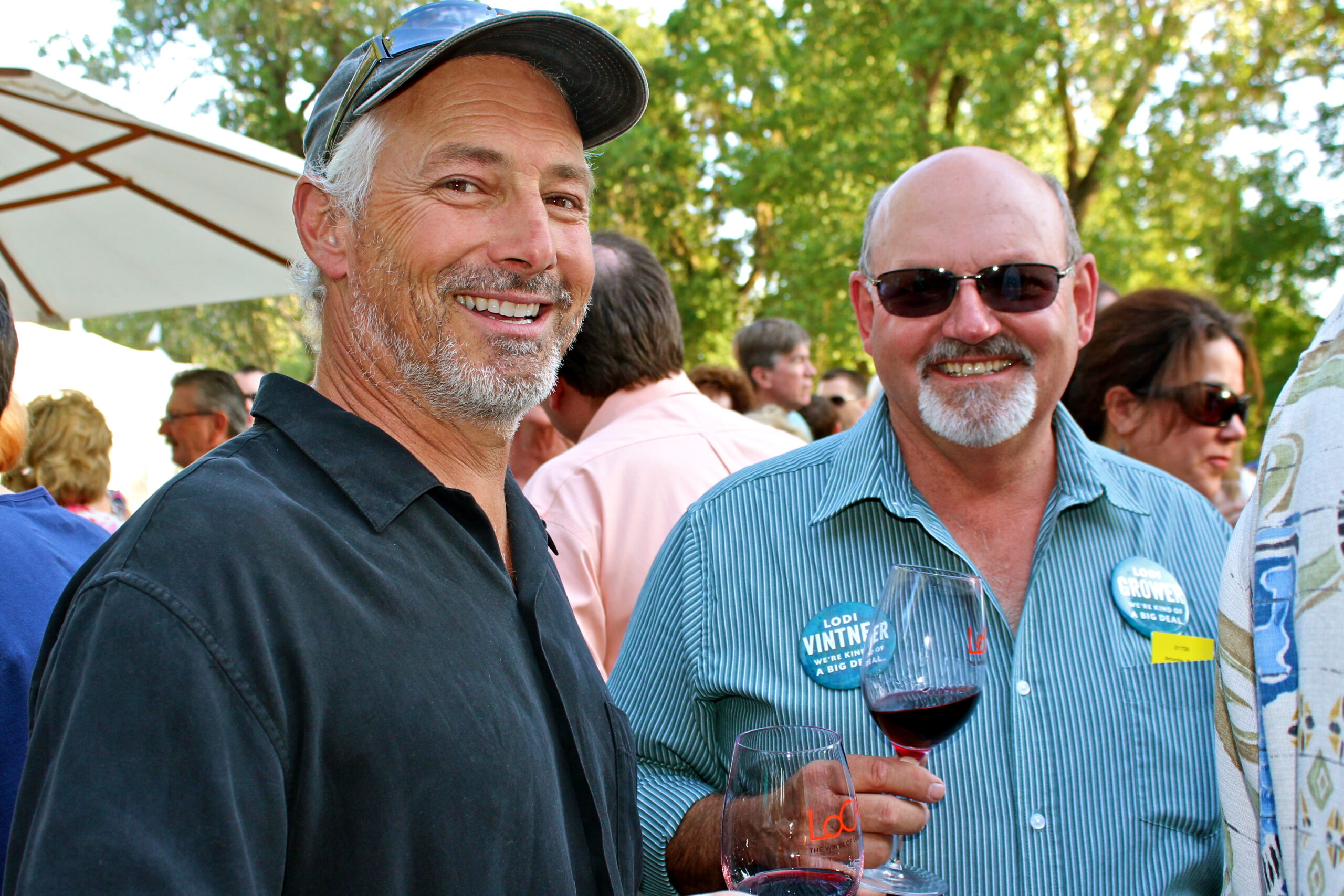 Two men enjoying wine at a Lodi Winegrape Commission event outdoors with trees and other attendees in the background.