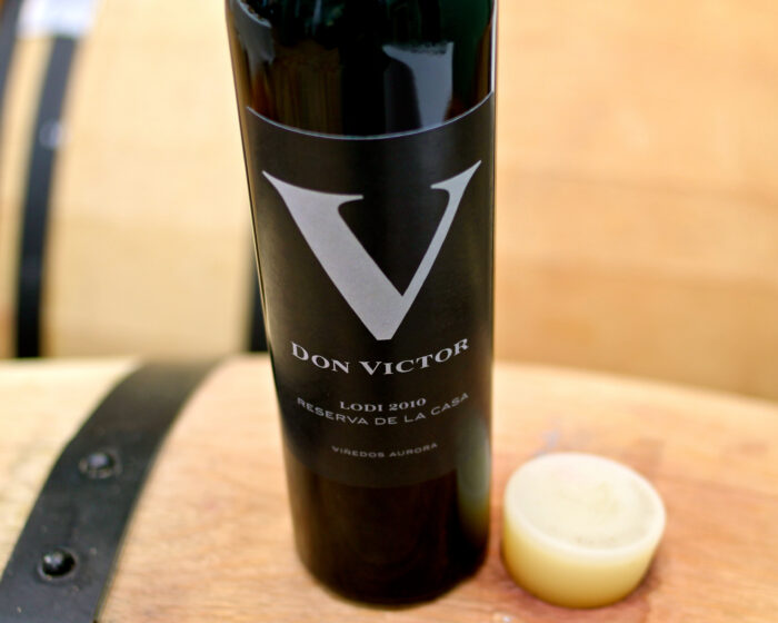 Lodi 2010 Don Victor wine bottle from the Lodi Winegrape Commission, on a wooden surface with a cheese wheel nearby.