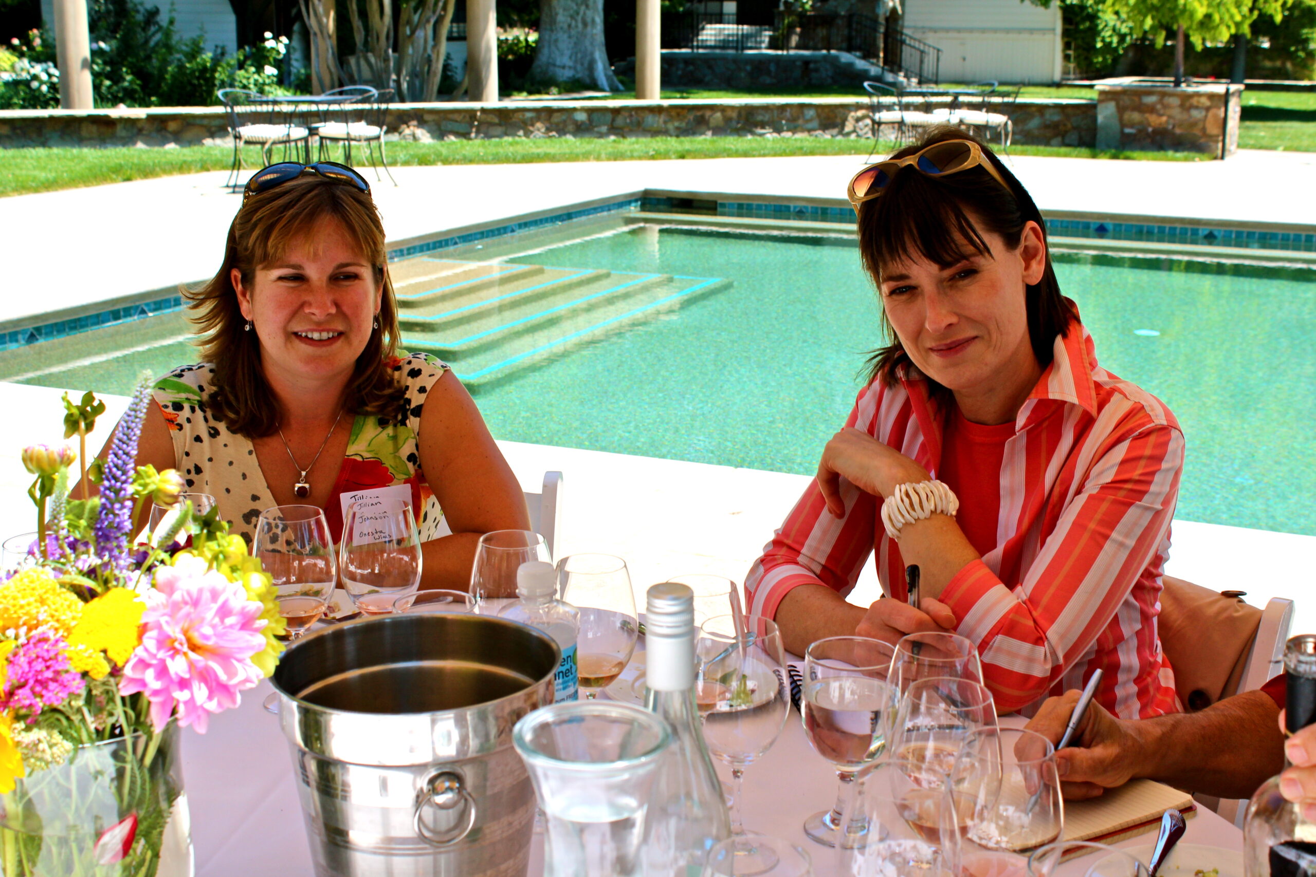 Two women enjoying a wine tasting event outdoors with a pool in the background.