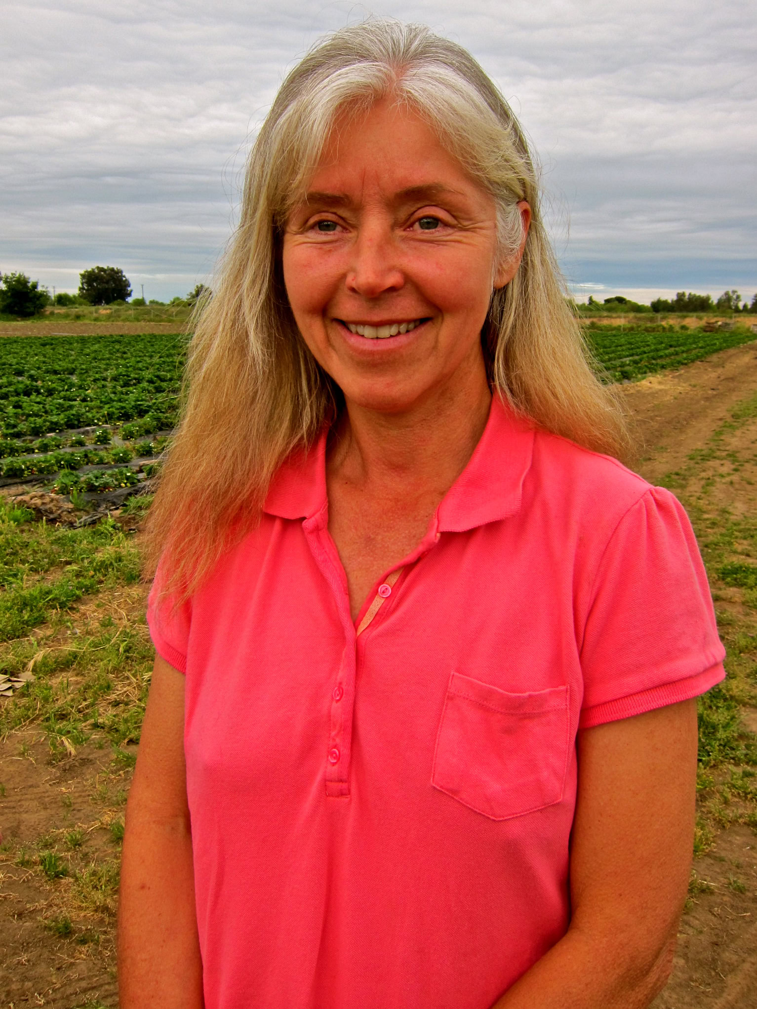 Smiling woman in a pink polo shirt standing in a vineyard with grapevines and cloudy sky.